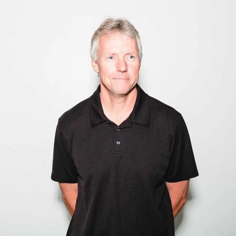 A middle-aged man with light hair and a beard, wearing a black polo shirt, standing against a plain white background.