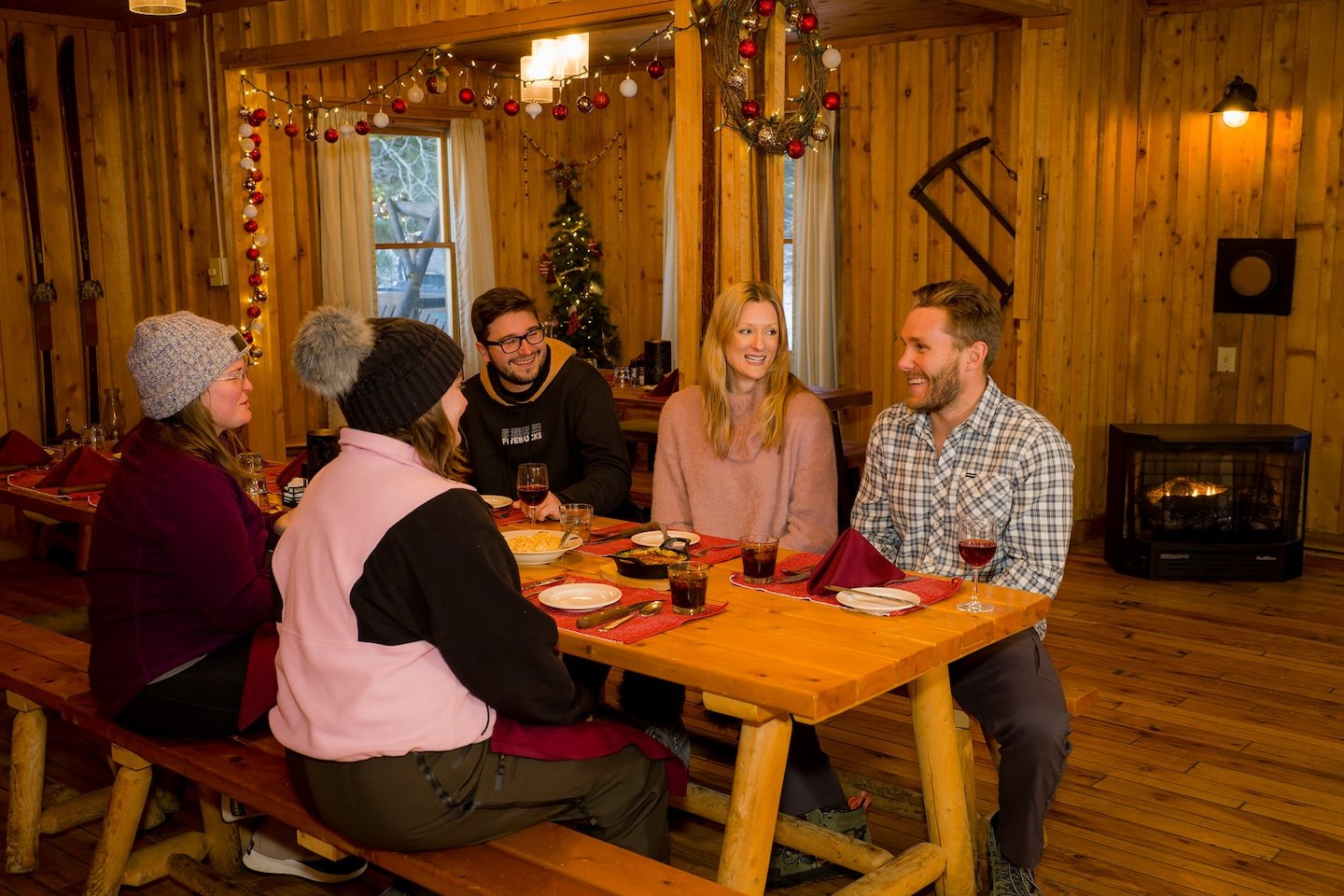 A group of five friends sitting around a wooden table in a cozy, wood-paneled room decorated for Christmas with string lights, ornaments, and a small Christmas tree, enjoying a meal and drinks, smiling and chatting.