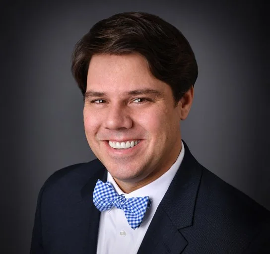 Professional portrait of a man wearing a dark suit and a blue and white checkered bow tie, smiling against a dark gray background.