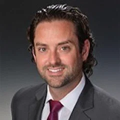 Professional headshot of a man with dark hair, beard, wearing a gray suit and red tie, smiling against a dark background.