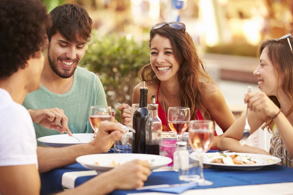 Four friends enjoying a meal and laughing together outdoors at a restaurant table with wine glasses.