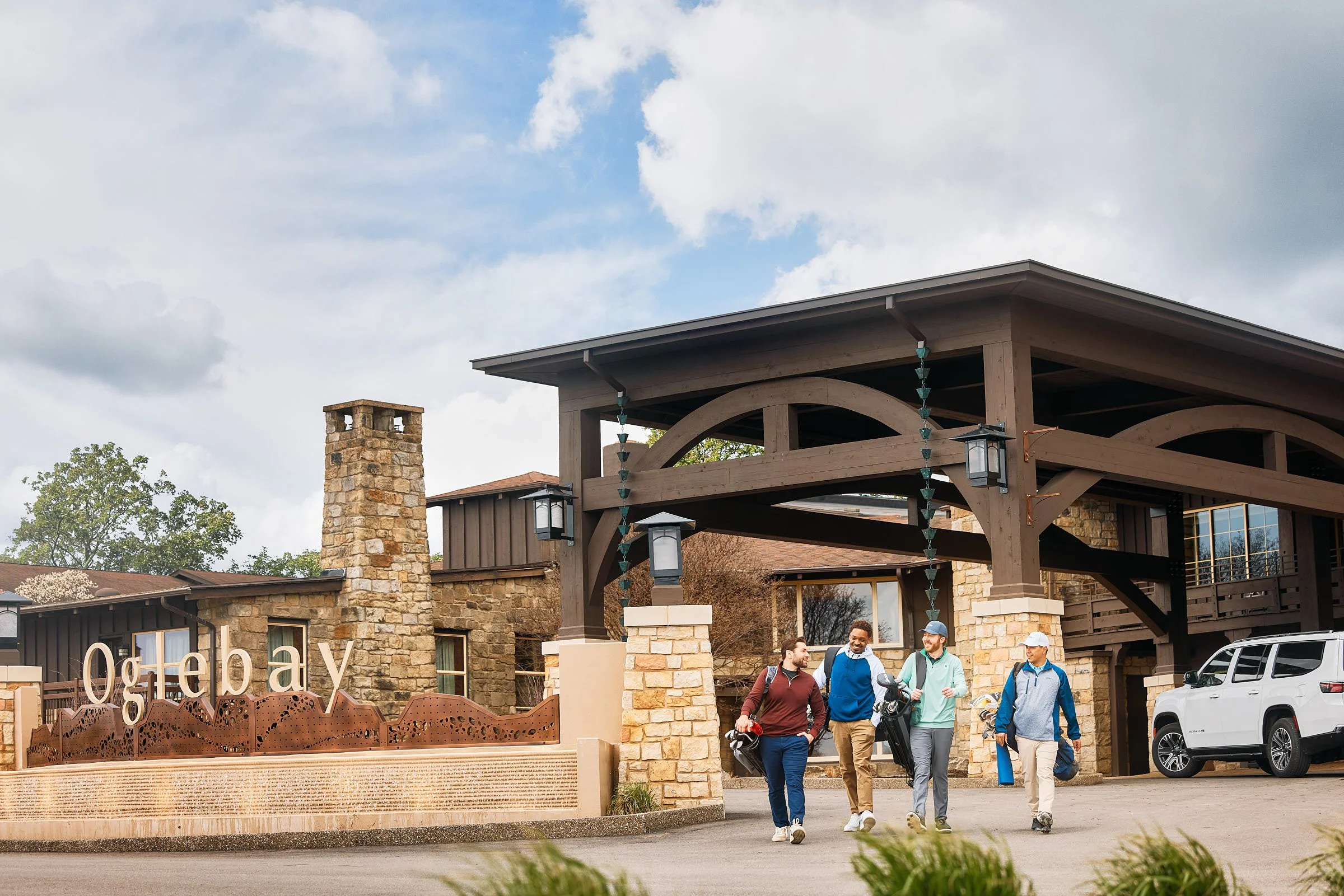 Group of four men walking out of a clubhouse or lodge, smiling and chatting, with a golf club bag. The building has stone and wood architecture with large lamps hanging from the roof. A golf cart is parked beside the building, and greenery is in the foreground.