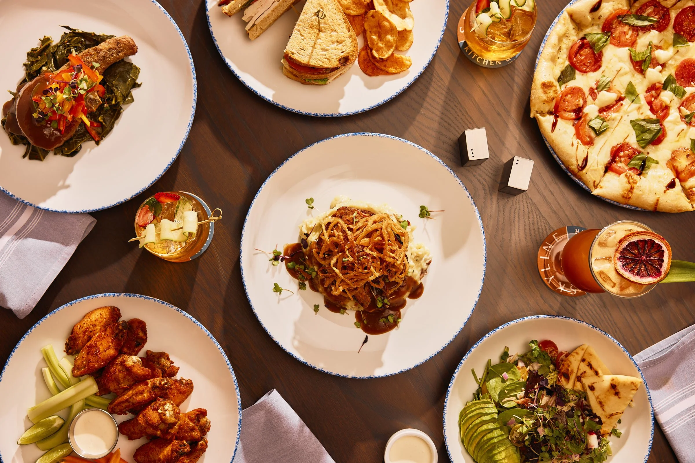 A table set with various dishes including a slice of pizza, a salad with avocado, fried chicken wings with celery and dipping sauce, a sandwich with chips, a plate of beef with mashed potatoes, a drink with a citrus slice, and two small glasses of drinks, all on white plates with a blue rim.