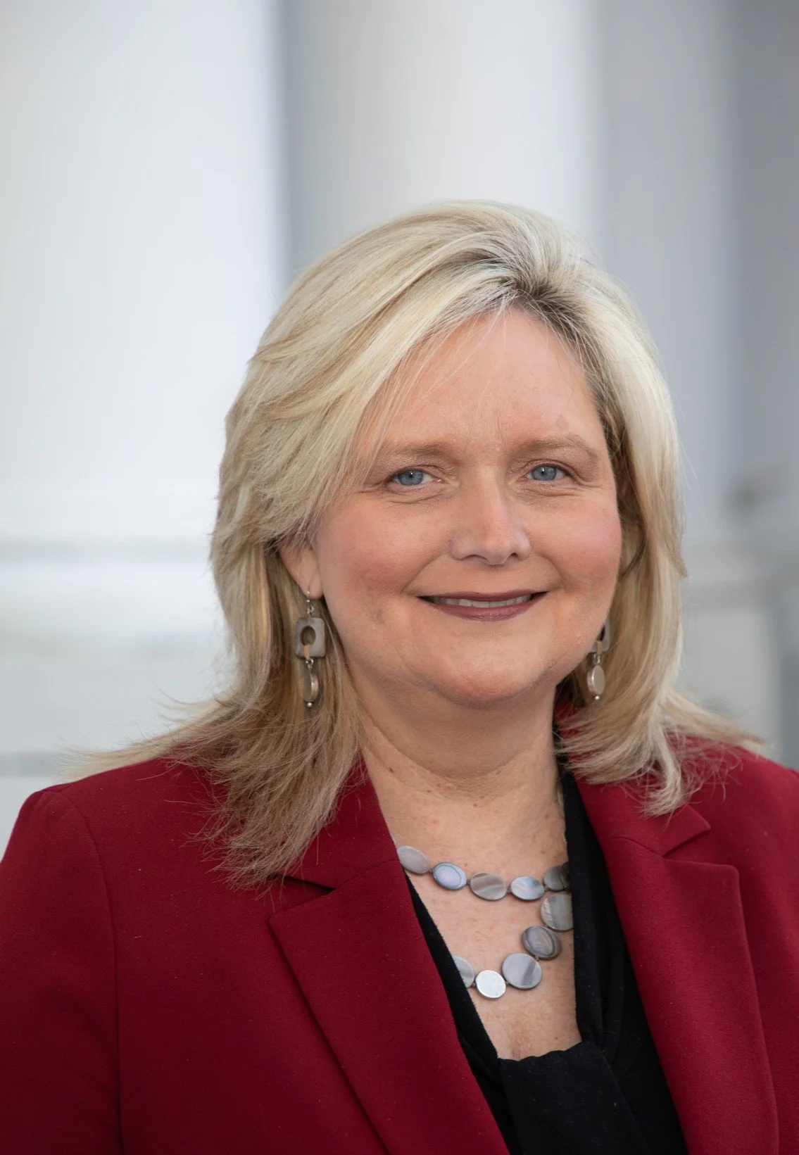 A woman with blonde hair, blue eyes, wearing a red blazer, black top, and jewelry including earrings and a necklace, smiling at the camera.