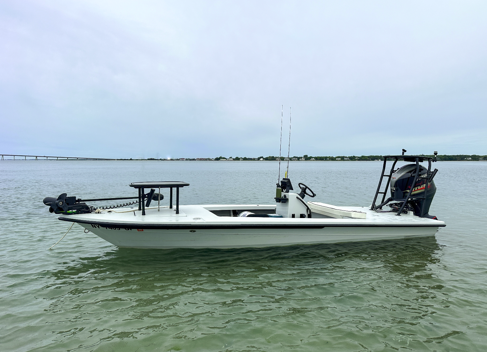 Small white fishing boat with a motor at the back, floating on calm water near a distant shoreline with houses and a bridge, under an overcast sky.
