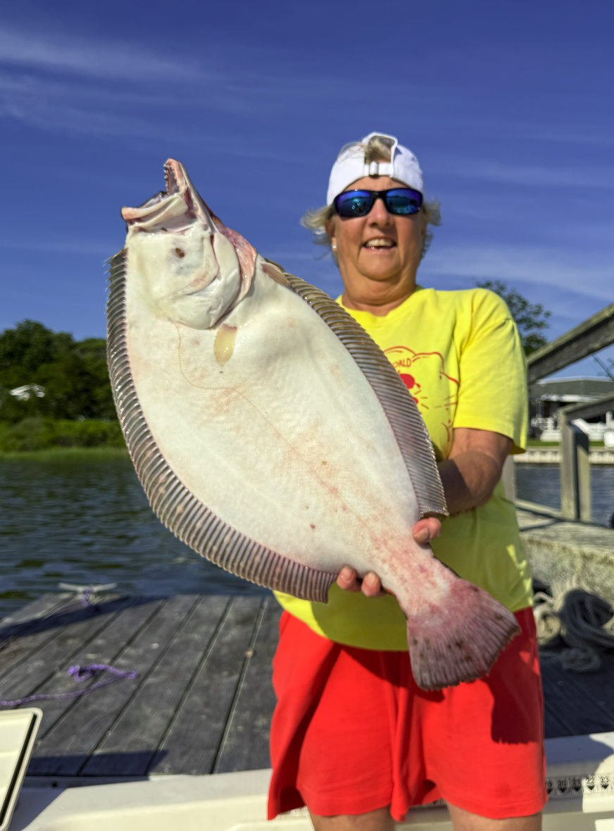 Smiling woman wearing sunglasses, a yellow T-shirt, and a white cap, holding a large flatfish with a light-colored underside. She is on a dock by the water with trees and a blue sky in the background.