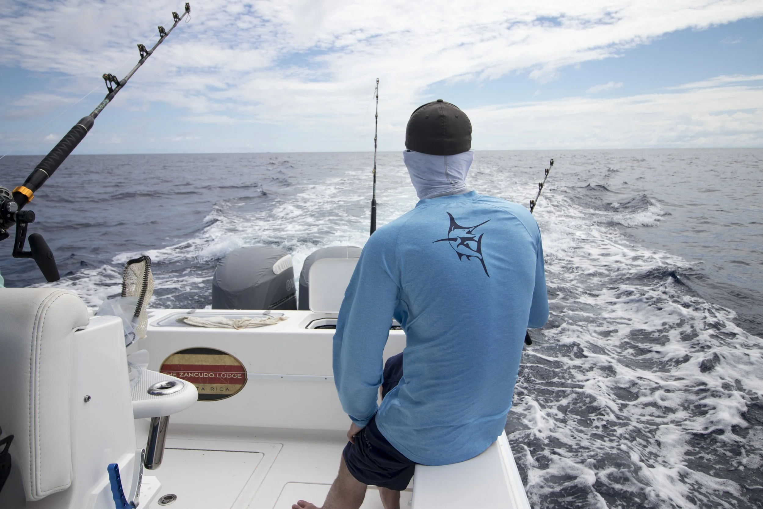 A person sitting at the back of a boat, fishing in the open ocean on a partly cloudy day.