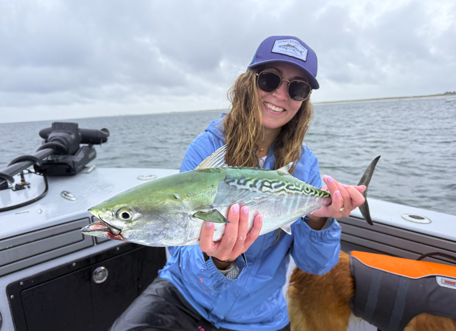 Woman in blue jacket and sunglasses holding a large fish on a boat with water and cloudy sky in the background.