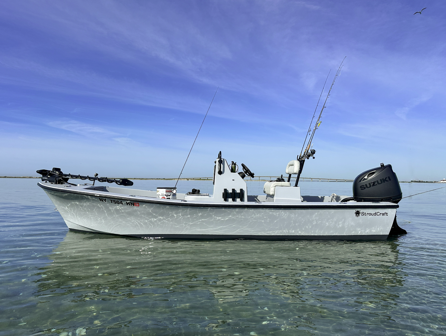A white fishing boat with fishing rods attached, floating on calm water under a blue sky with some clouds and a bird flying overhead.