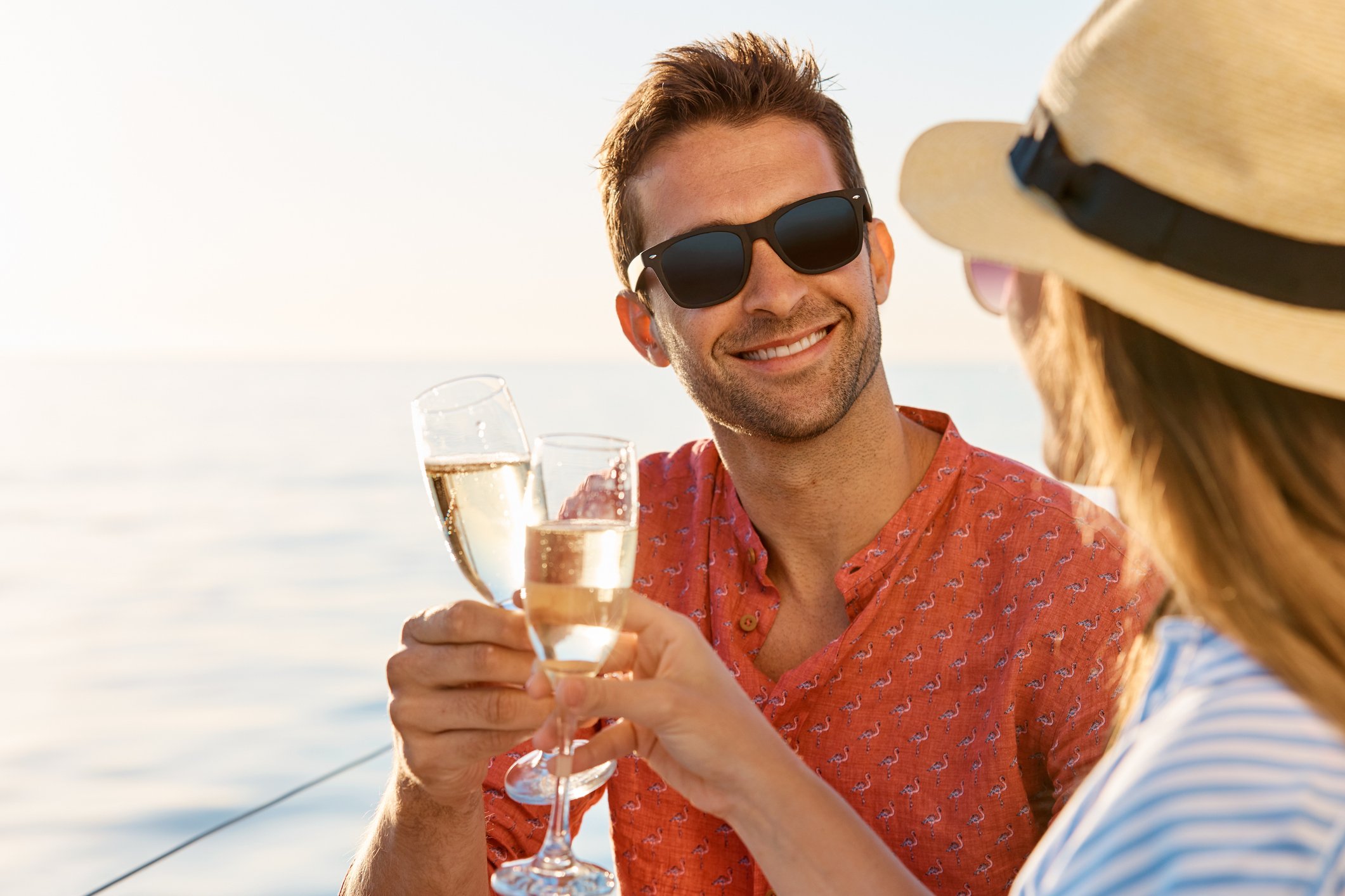 A man with brown hair, wearing sunglasses and a patterned orange shirt, smiling and holding a glass of champagne, toasting a woman with a wide-brimmed hat and sunglasses, on a boat during sunset or sunrise by the water.