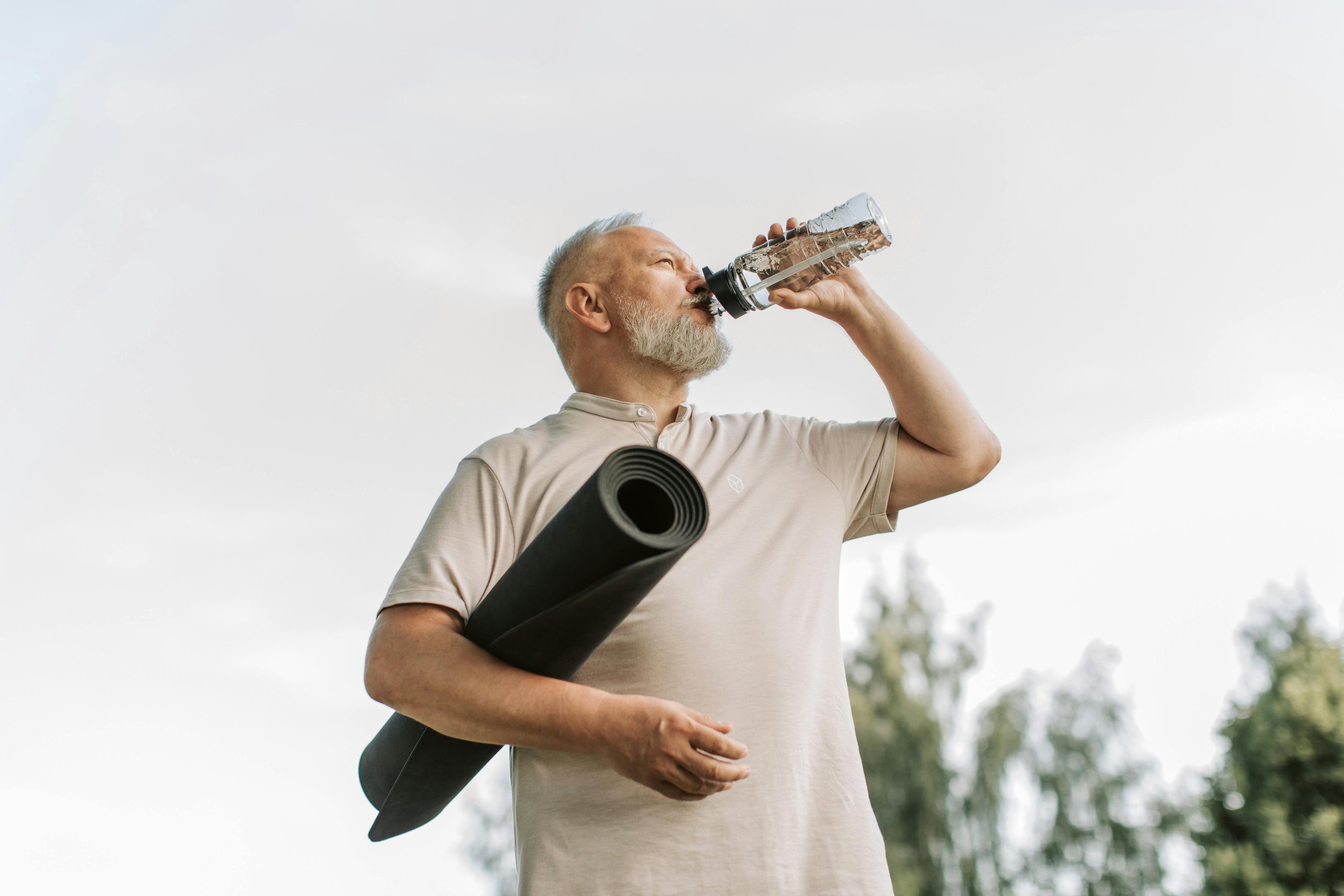 Un homme d'âge moyen avec une barbe grise, portant un t-shirt beige, buvant de l'eau d'une bouteille en verre, tenant un tapis de yoga noir, en extérieur avec des arbres en arrière-plan.