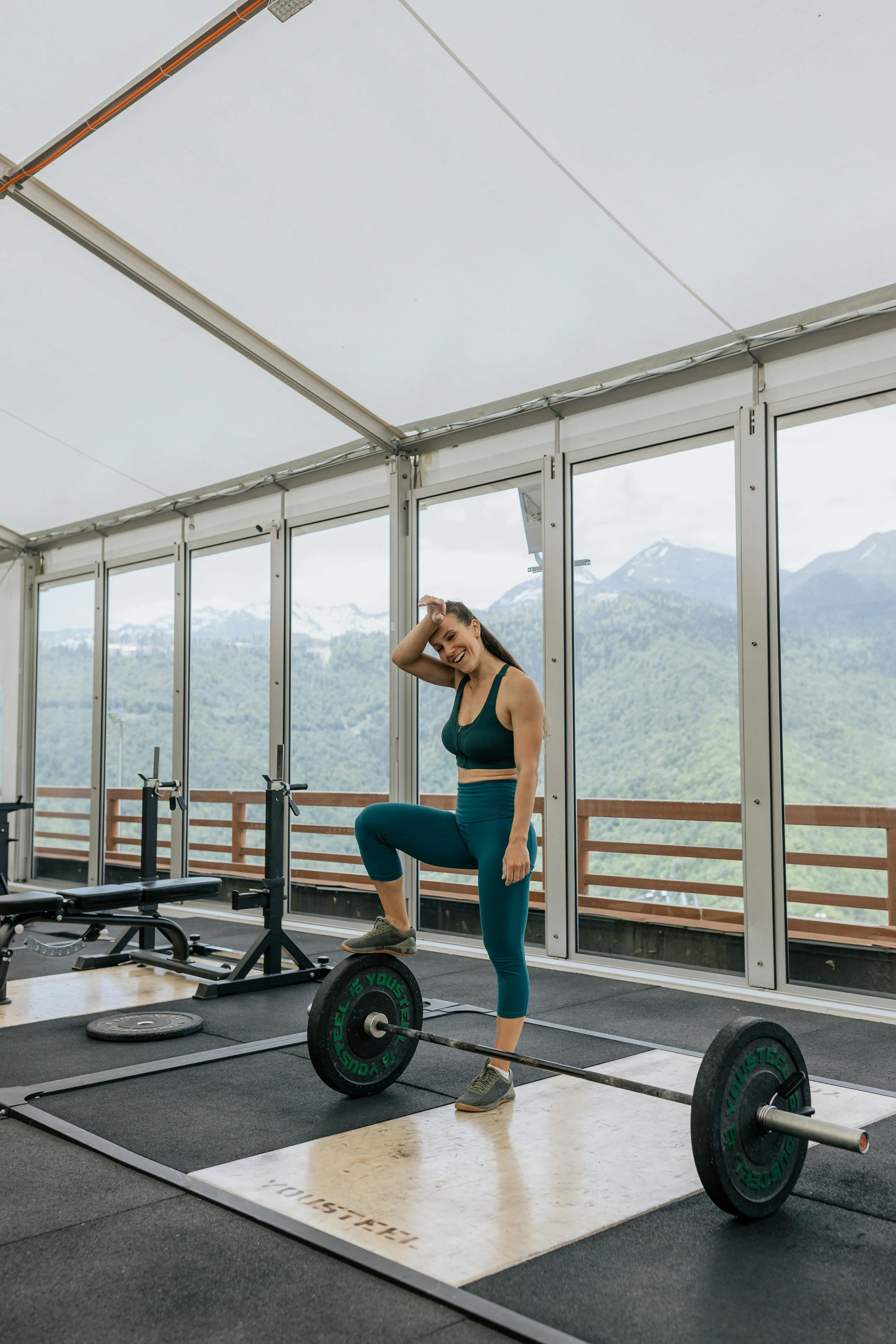 Une femme en tenue de sport effectuant un entraînement de musculation avec un poids de barre dans une salle de sport, avec des montagnes en arrière-plan visible à travers de grandes fenêtres.