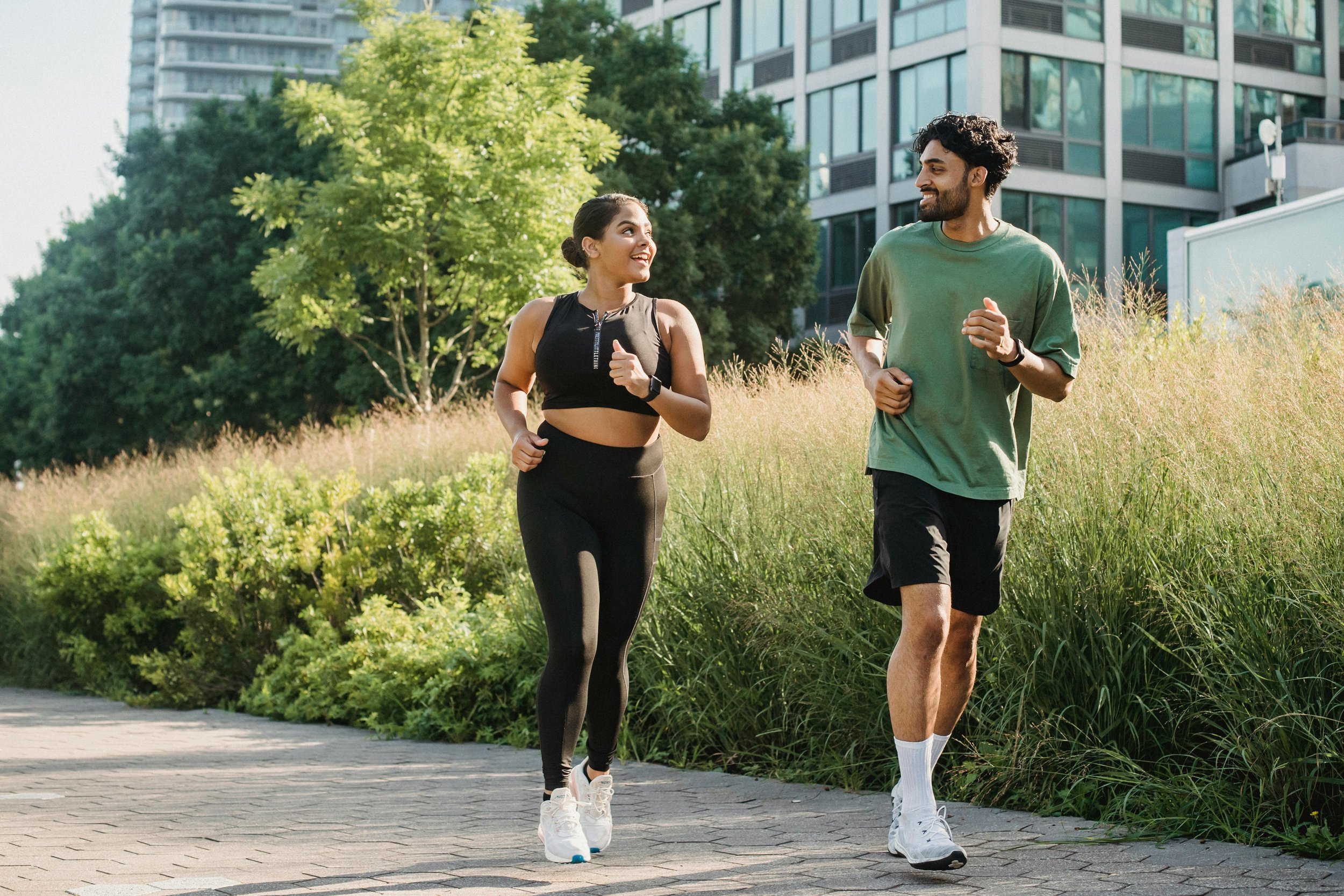 Deux personnes faisant du jogging dans un parc urbain, entourées d'arbres et de bâtiments modernes. La femme porte un pantalon de sport noir et un haut de sport noir, le homme porte un t-shirt vert et un short noir.