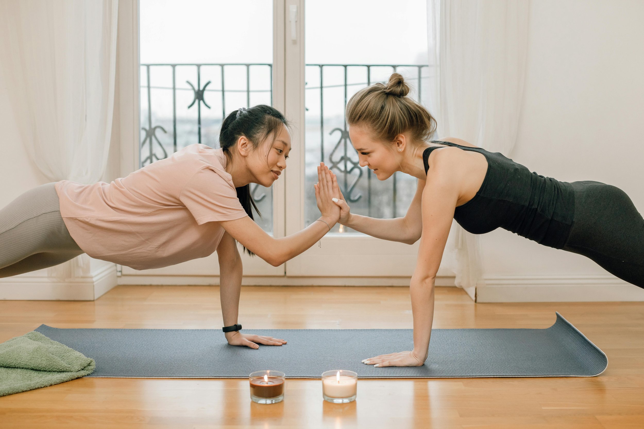 Deux femmes faisant du yoga en équilibre sur leurs mains, se touchant paume contre paume, dans une pièce lumineuse avec un balcon en arrière-plan.
