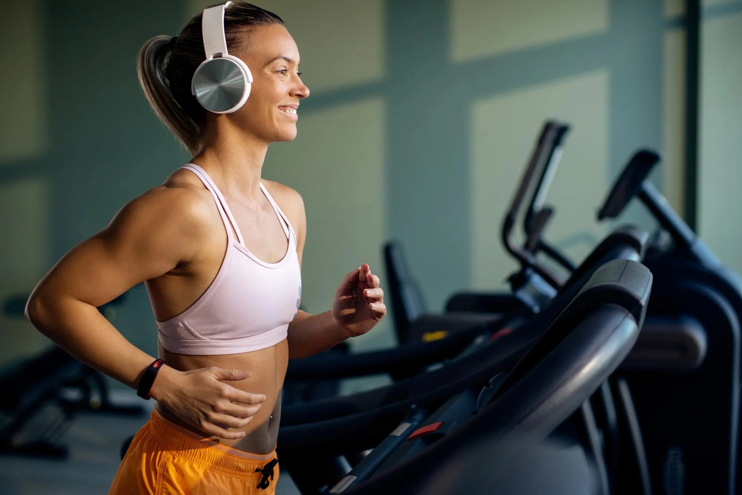 Femme en salle de sport, portant un casque audio, courant sur un tapis de course.