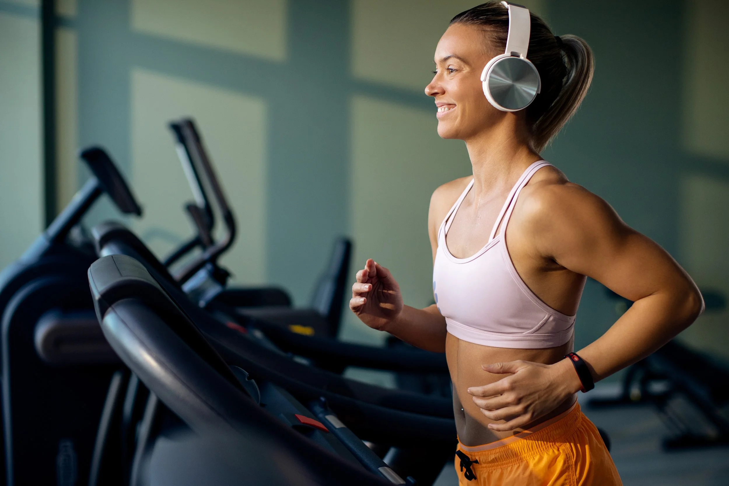 Femme souriante portant des écouteurs, en train de courir sur un tapis de course dans une salle de sport