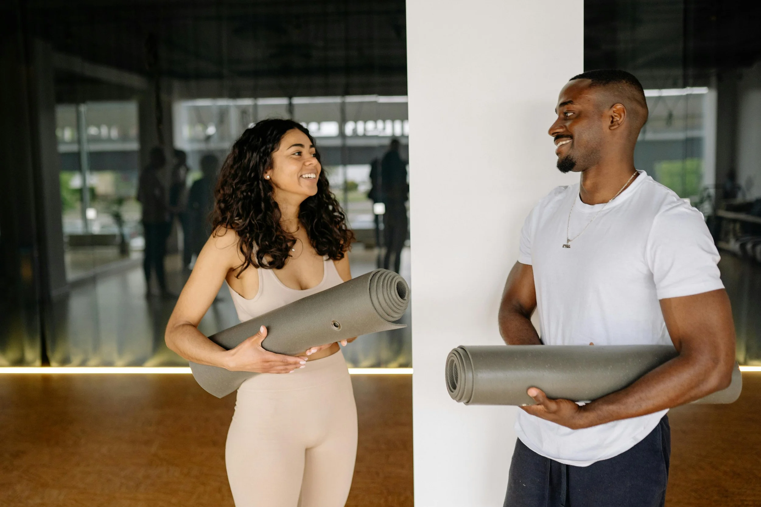 Une femme et un homme souriants tenant des tapis de yoga dans une salle de sport. La femme a les cheveux bouclés et porte des vêtements de sport beige. L'homme a les cheveux courts et porte un t-shirt blanc et un collier. Ils semblent discuter avant une séance d'entraînement.
