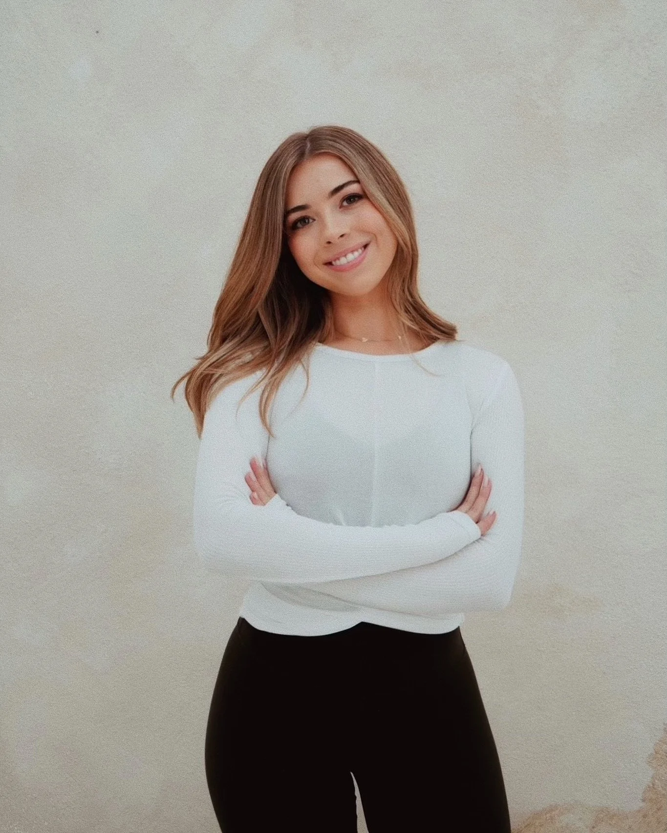 A woman with light brown hair, smiling, wearing a white long-sleeve top and black pants, standing against a plain off-white background.