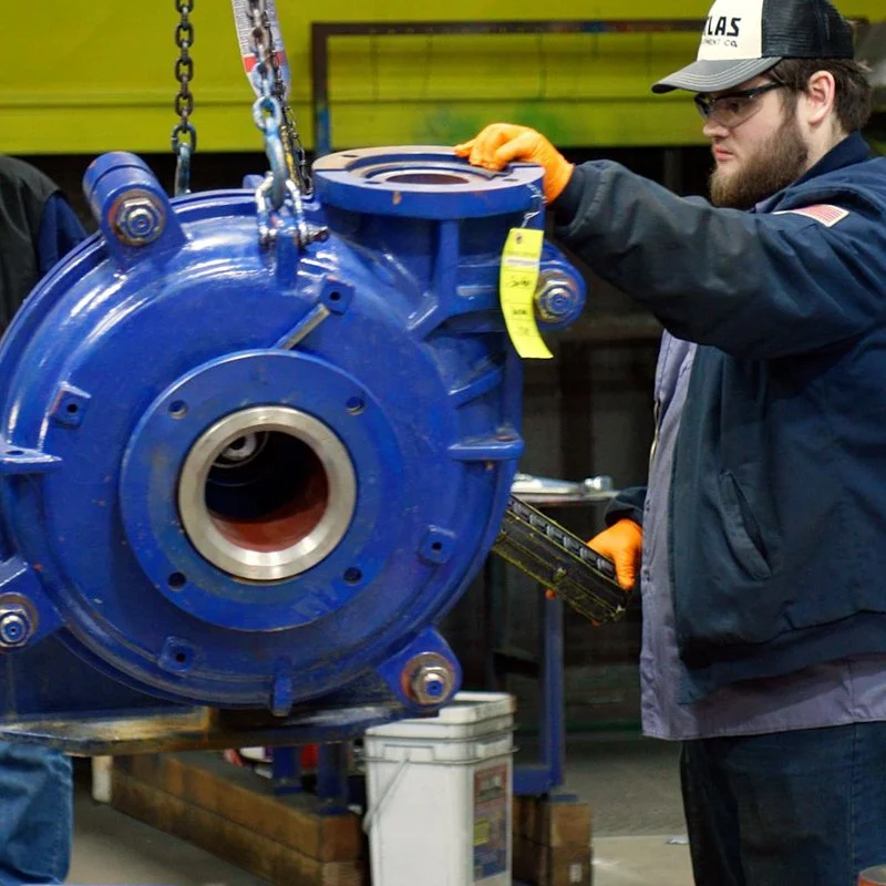 A man wearing safety gloves and glasses inspecting a large blue mechanical part in an industrial setting.