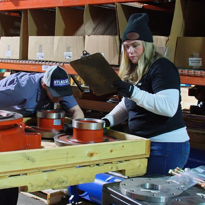 Two workers in a warehouse, inspecting large orange metal components on a wooden workbench. Shelves with boxes are visible in the background.