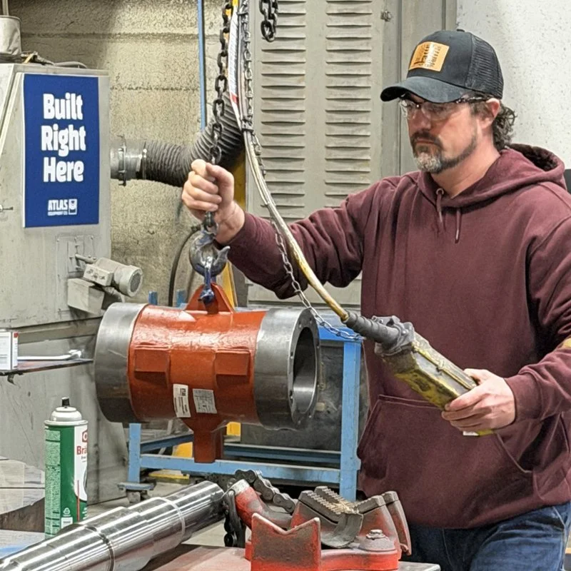 A man wearing safety glasses and a cap working with a hoist on a large metal machine part in an industrial setting.