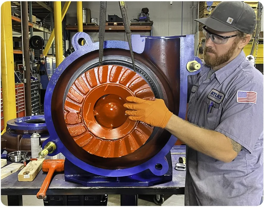A worker wearing safety glasses, gloves, a gray uniform, and a cap inspecting a large, rust-colored mechanical component in a workshop setting.