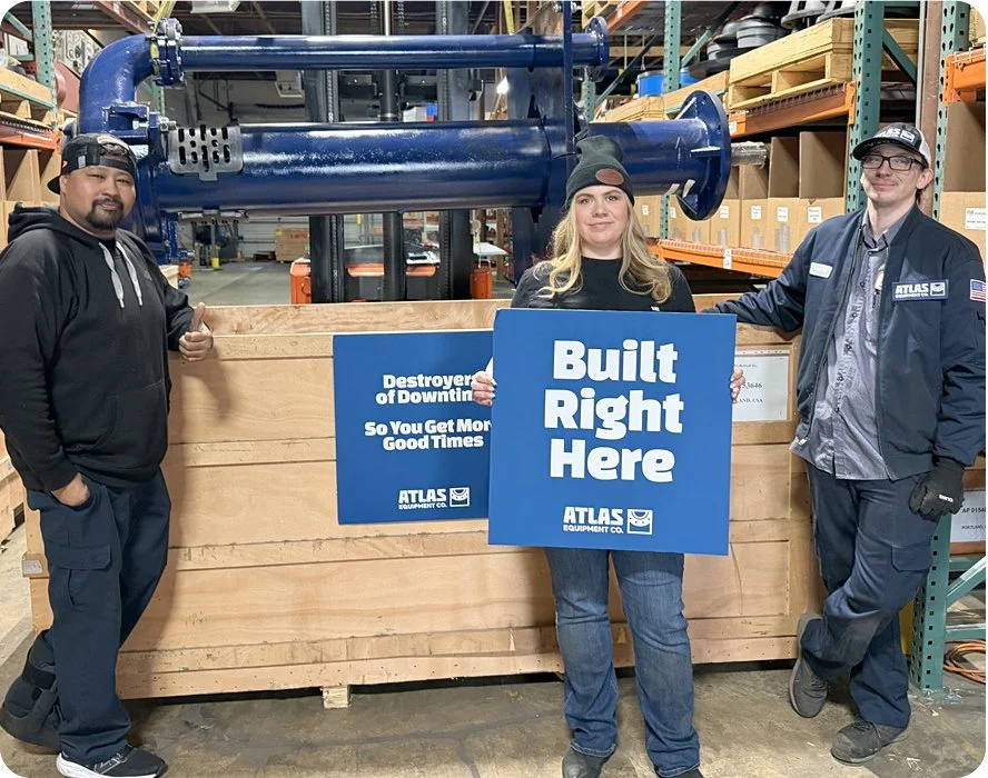 Three people standing in a warehouse near a large blue industrial pipe, holding blue signs with white text that read 'Built Right Here' and 'Destroyer of Downtowns So You Get More Good Times.'