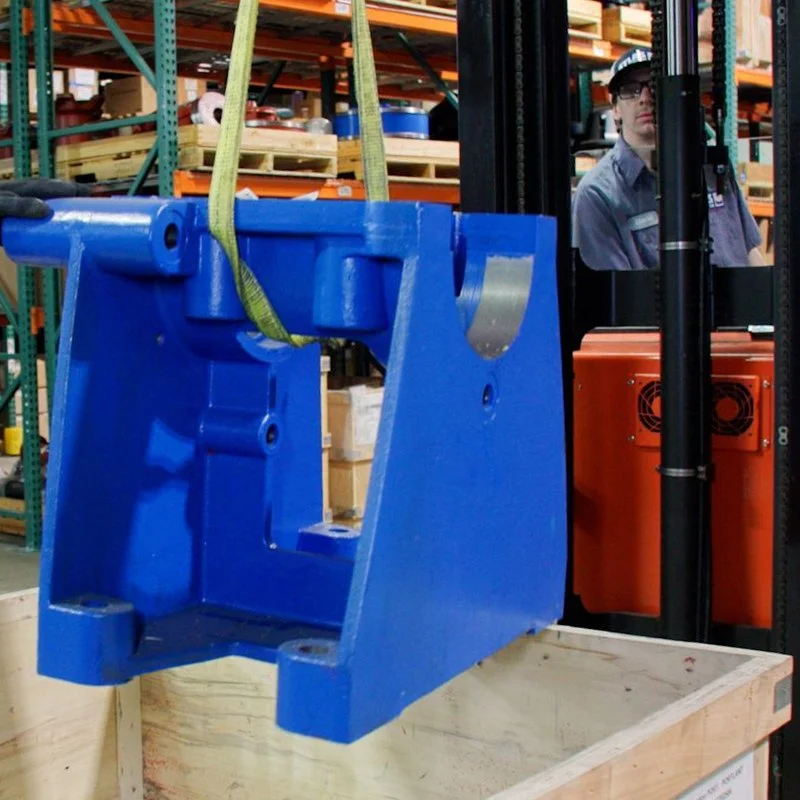 Close-up of a blue forklift attachment or component being lifted in a warehouse with shelves, boxes, and a worker wearing safety glasses and a uniform in the background.