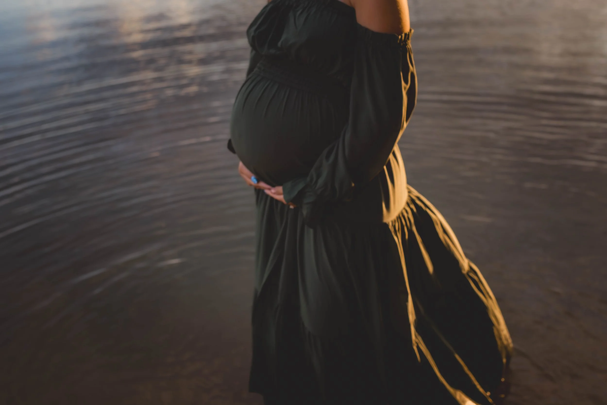 Pregnant woman standing in water, holding her belly with both hands, during sunset or sunrise.