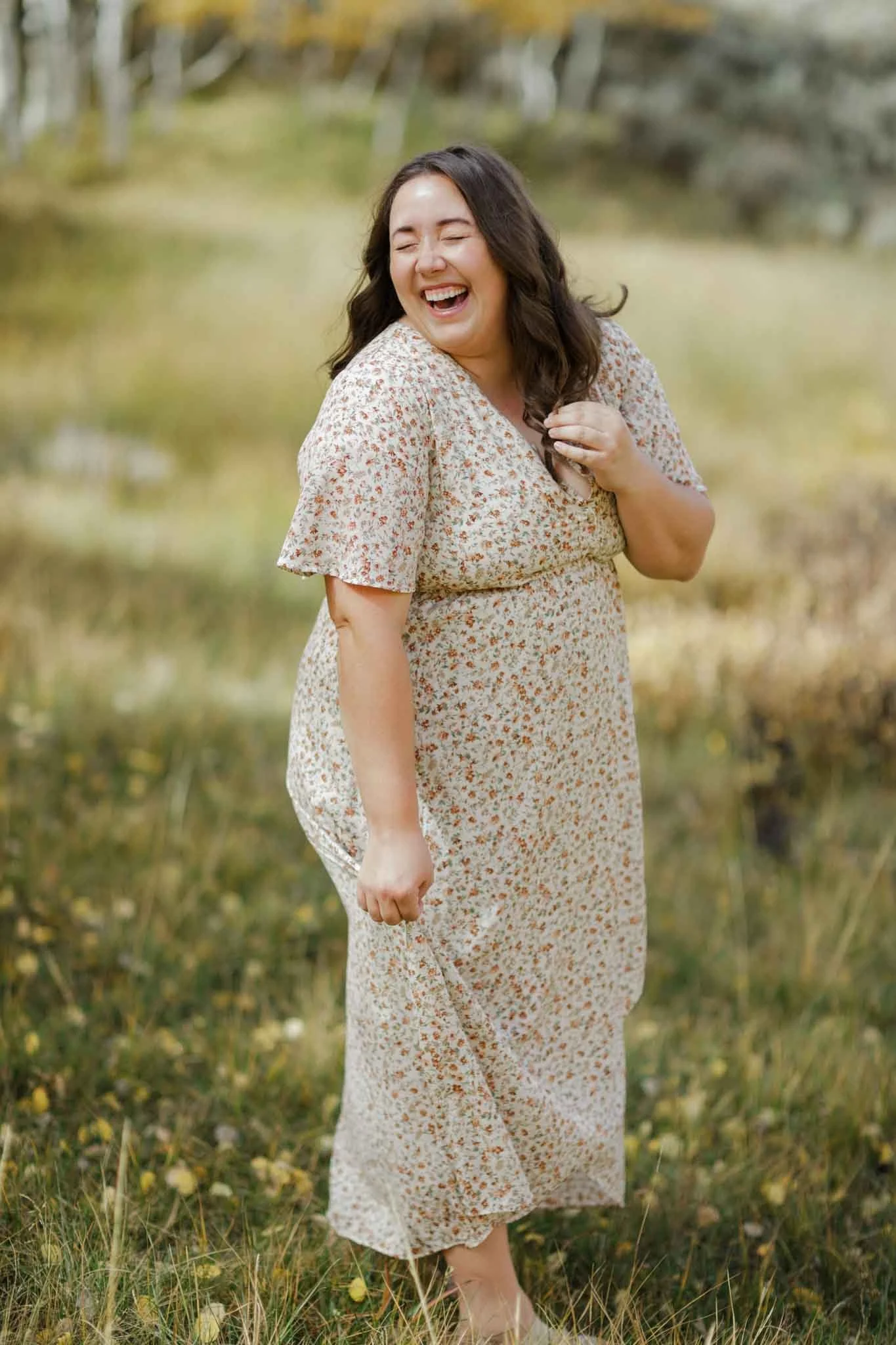 A woman with long dark hair, wearing a floral dress, laughing outdoors in a grassy area with trees in the background.