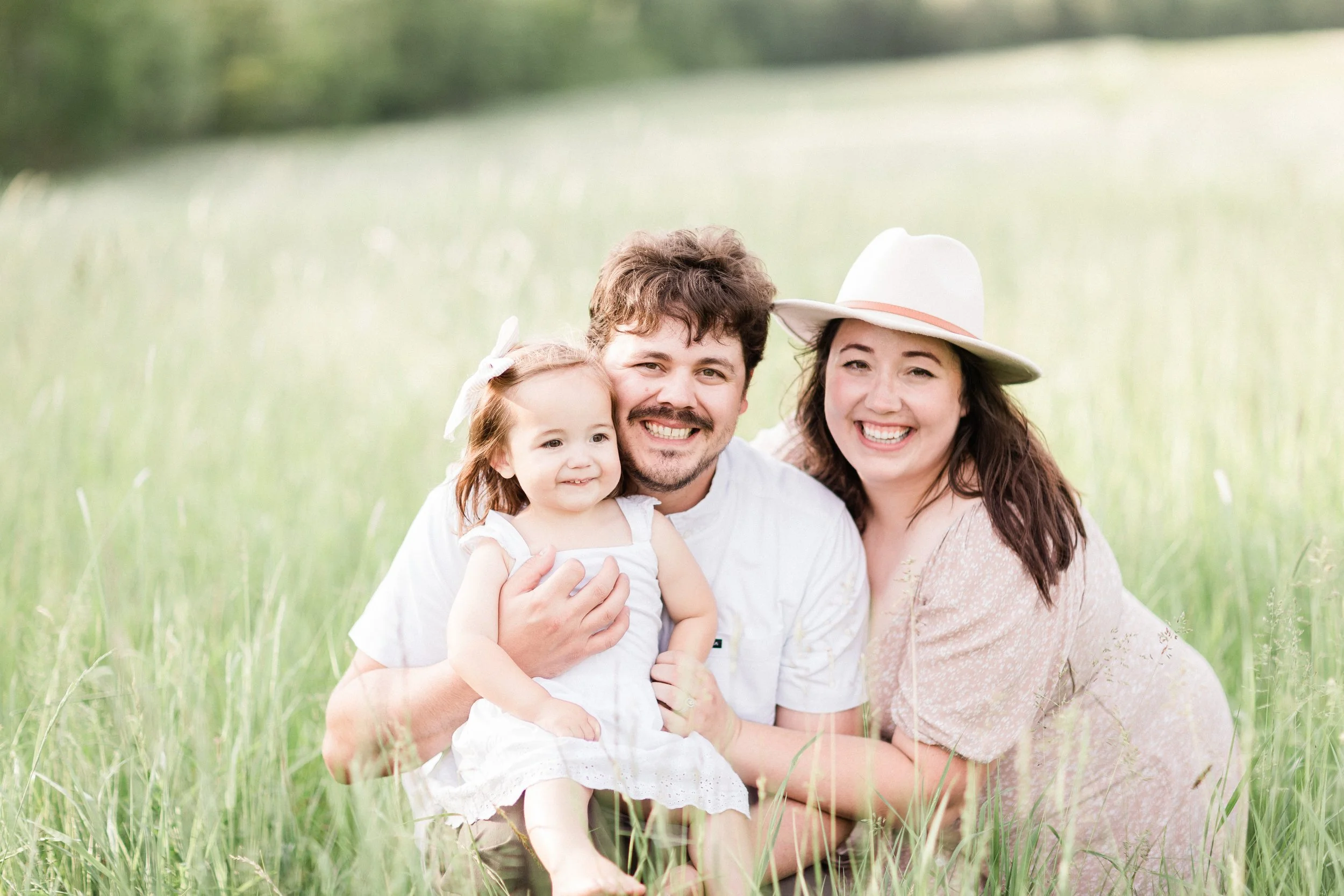 A happy family of three smiling together outdoors in a grassy field, including a man, a woman, and a young girl in white dresses, with the woman wearing a wide-brimmed hat.