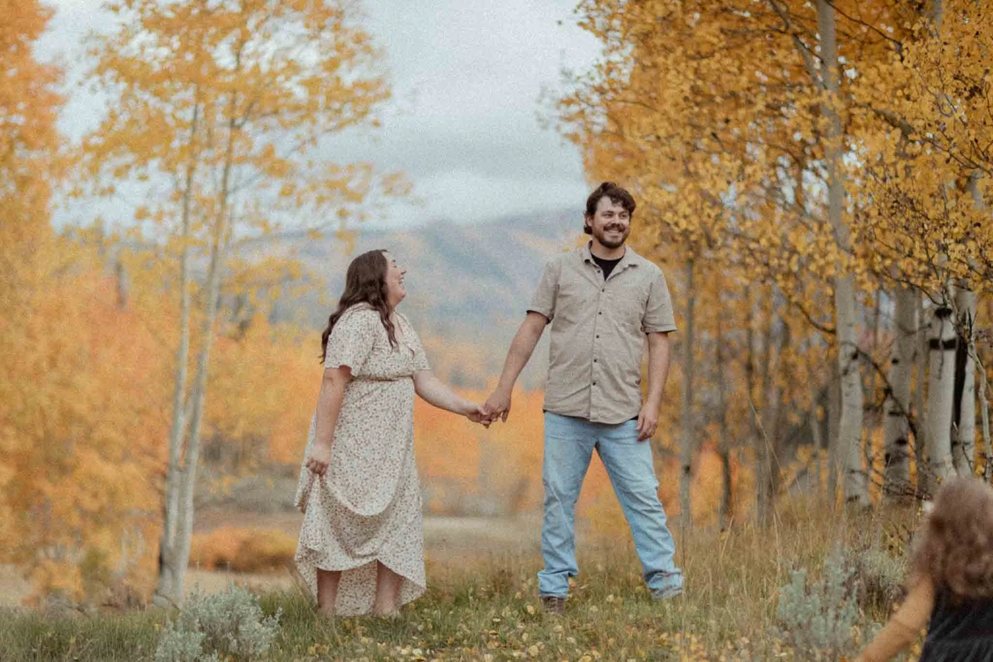 A couple holding hands outdoors among fall trees with orange leaves. The woman is wearing a long dress, and the man is in a tan shirt and jeans. The woman is looking at the man, who is smiling and looking away. There is a child with curly hair partially visible in the foreground.