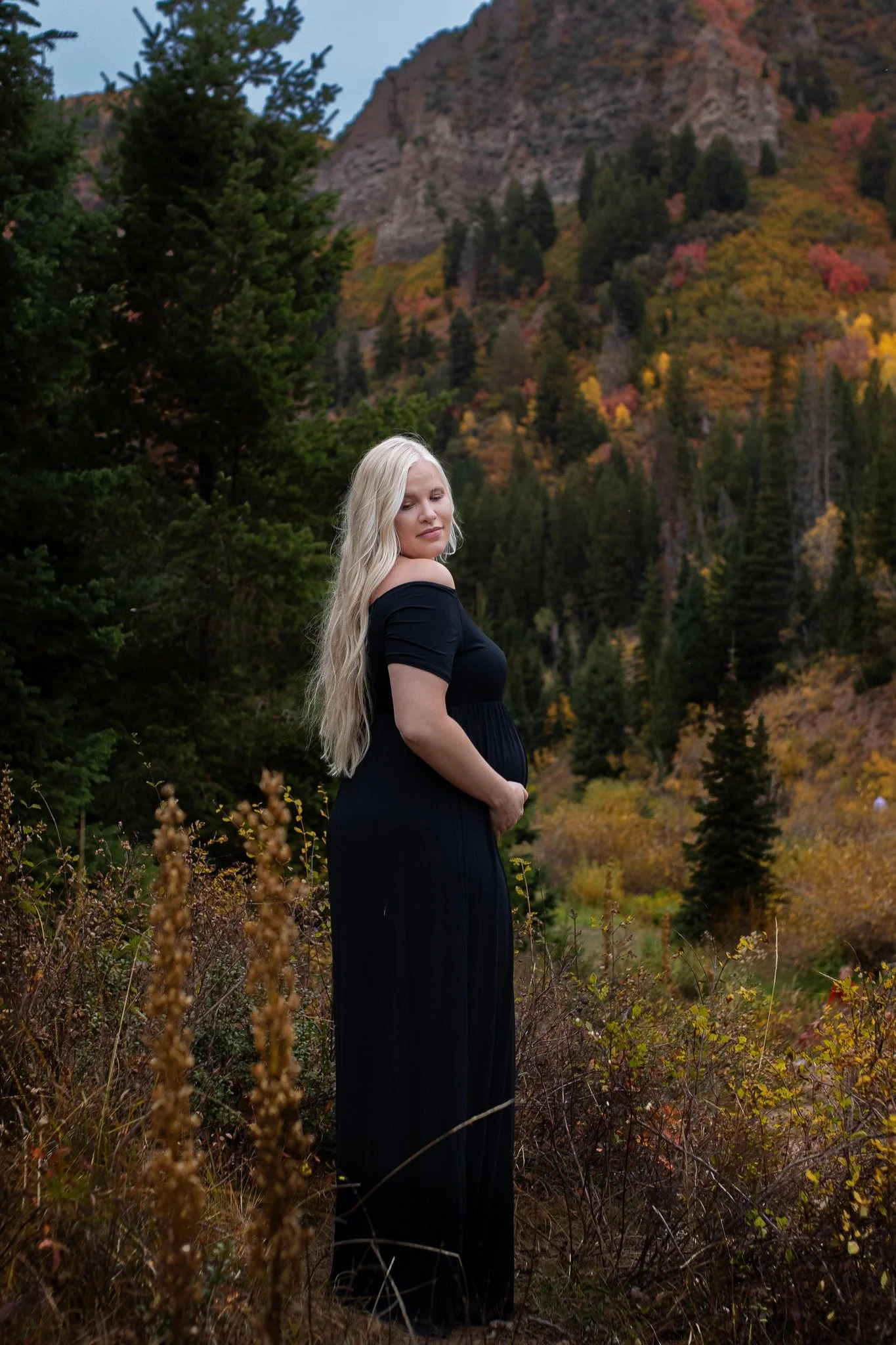 A pregnant woman in a black dress standing in a natural landscape with trees and mountains in the background during autumn.