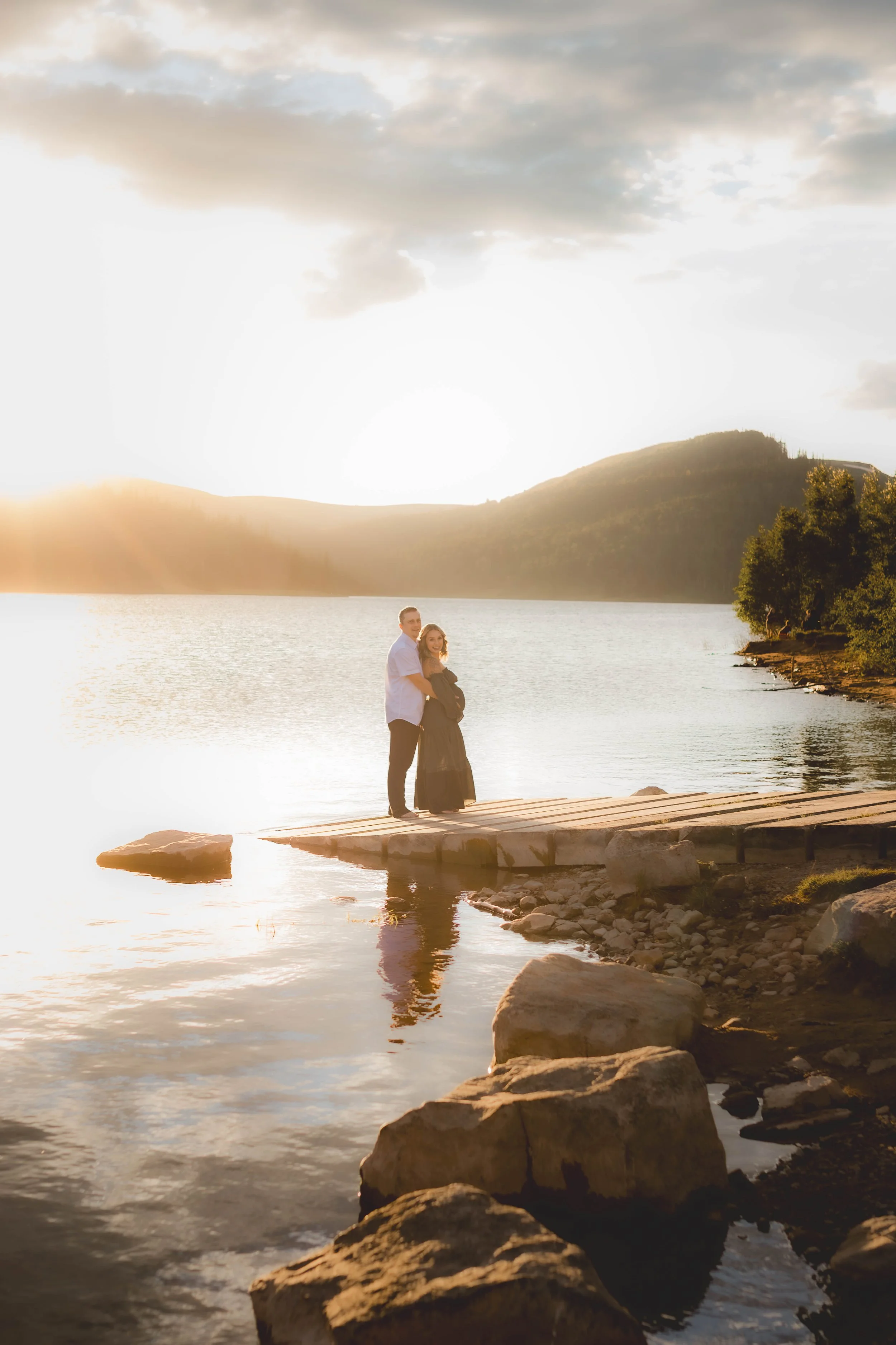 A couple standing on a wooden dock by a lake during sunset, with mountains and trees in the background.