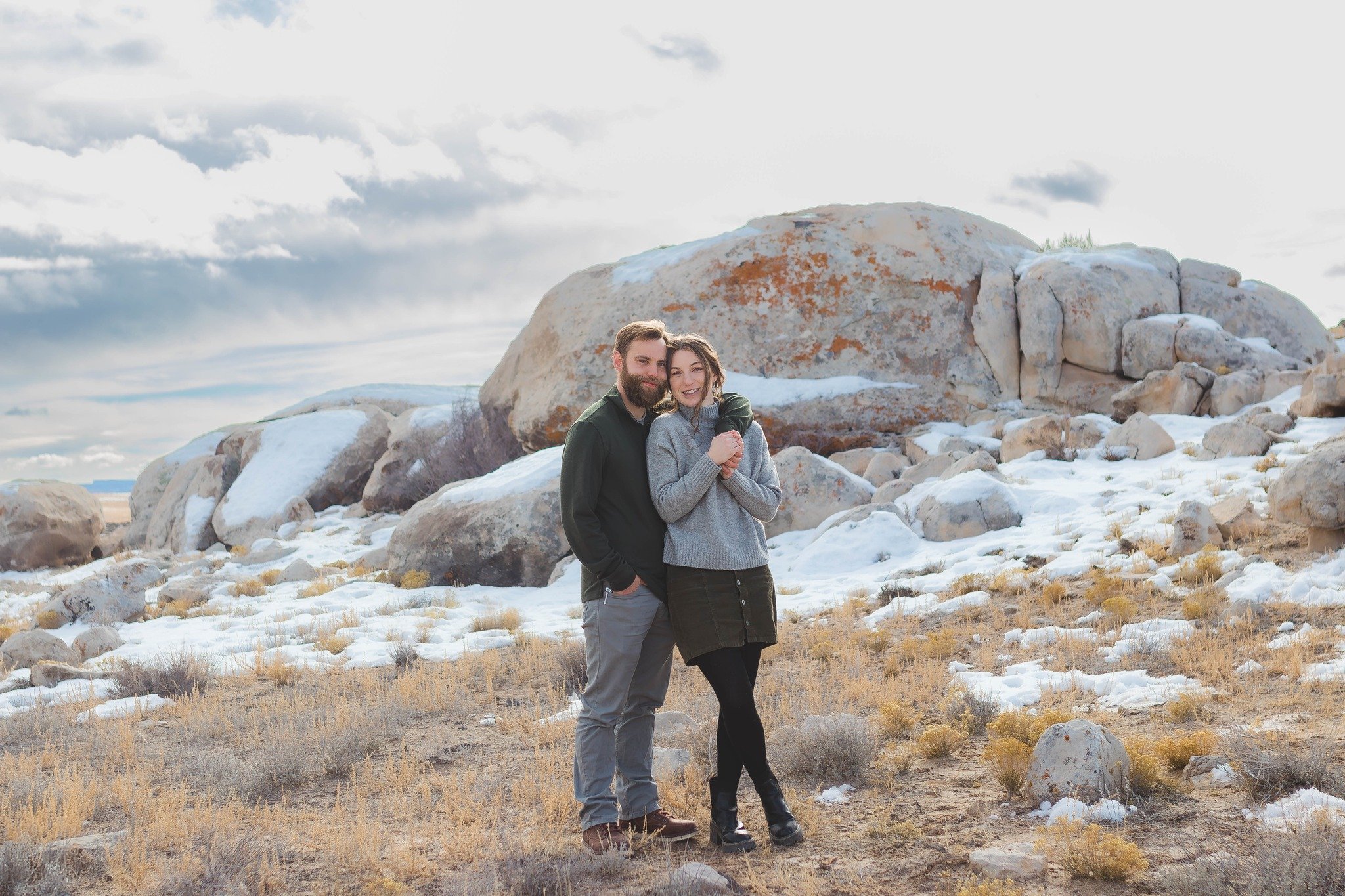 A couple stands in a rocky, snowy landscape, smiling and embracing each other.