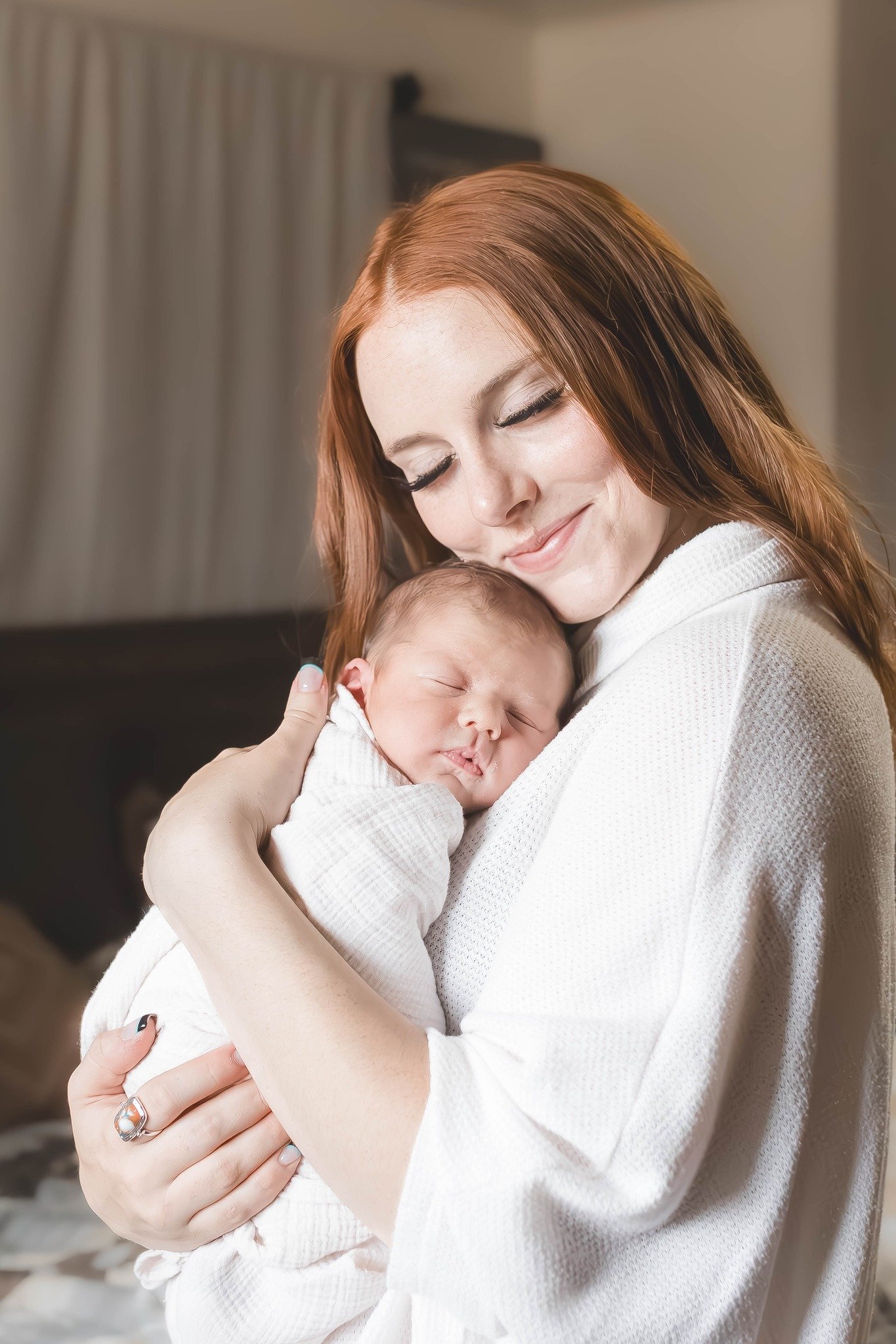 A woman with red hair holding a sleeping baby wrapped in a white blanket, smiling gently.