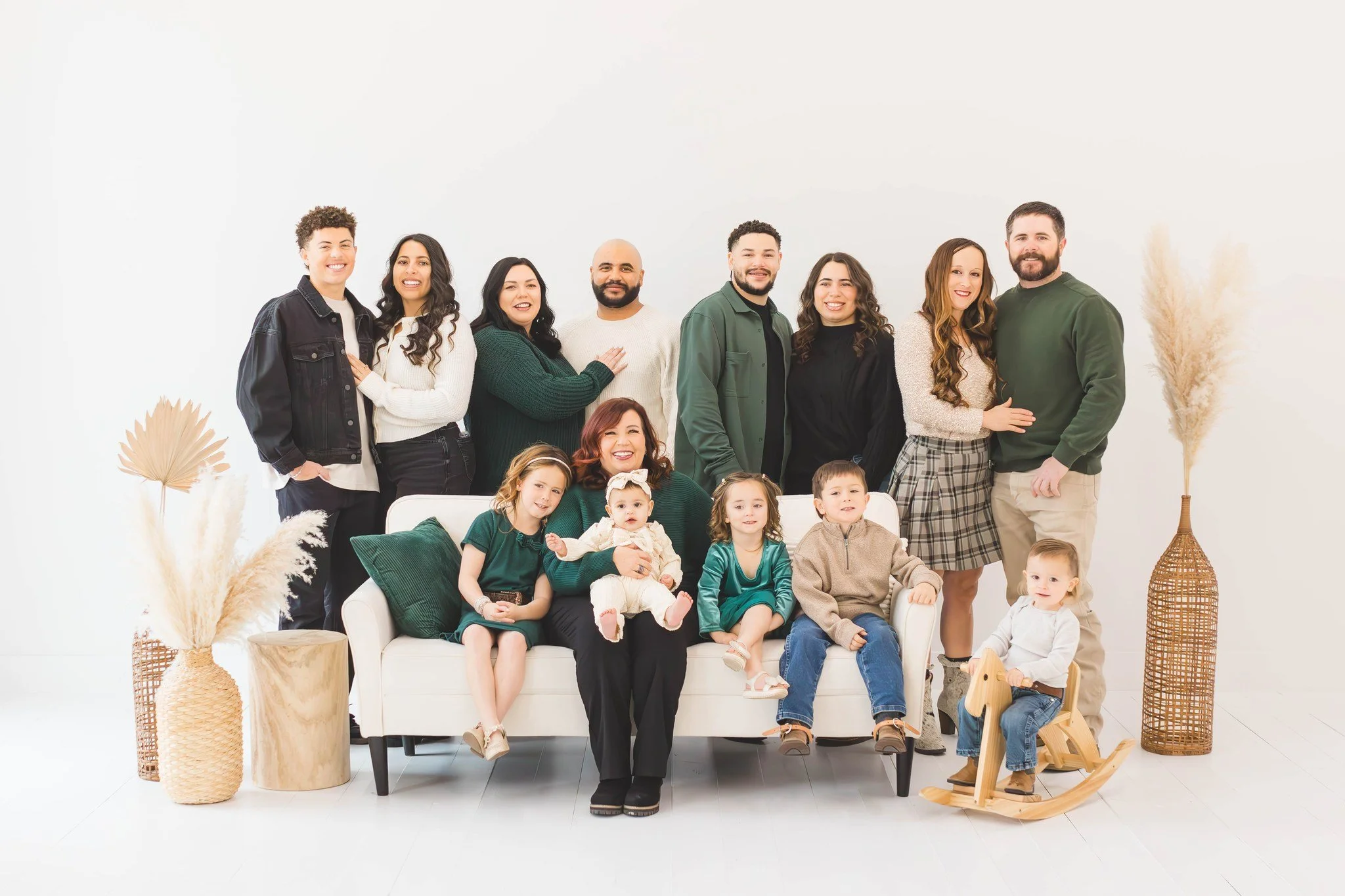 A large multigenerational family group portrait in a bright, minimalistic room with white walls. The family members, dressed in casual, warm clothing, are gathered around a white sofa with some sitting on it and others standing behind. Children are seated on the sofa and one is on a wooden rocking horse. Decorative dried plants in woven baskets are placed on either side of the group.