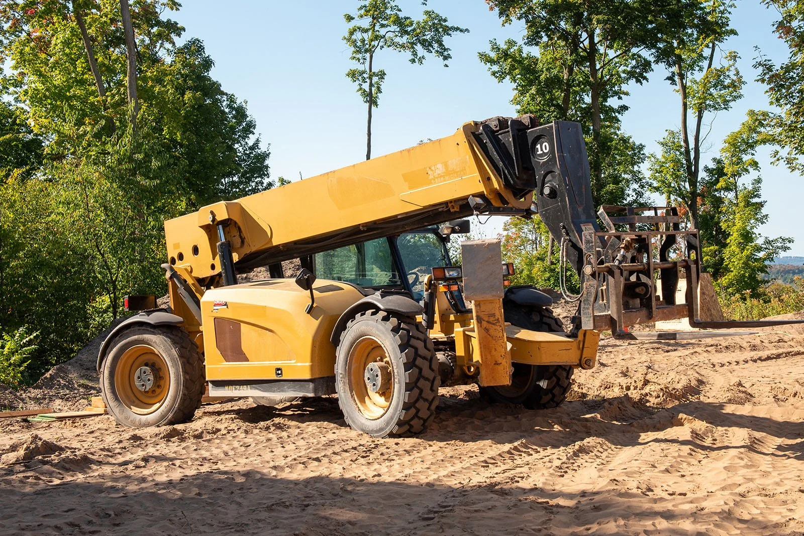 Yellow construction telehandler on a sandy construction site surrounded by trees.