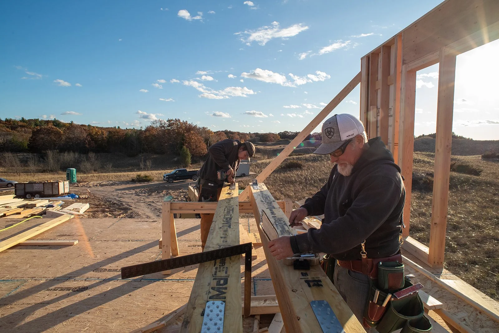 Two people working together on a construction site, nailing wooden beams to a wall, with a red ladder nearby.
