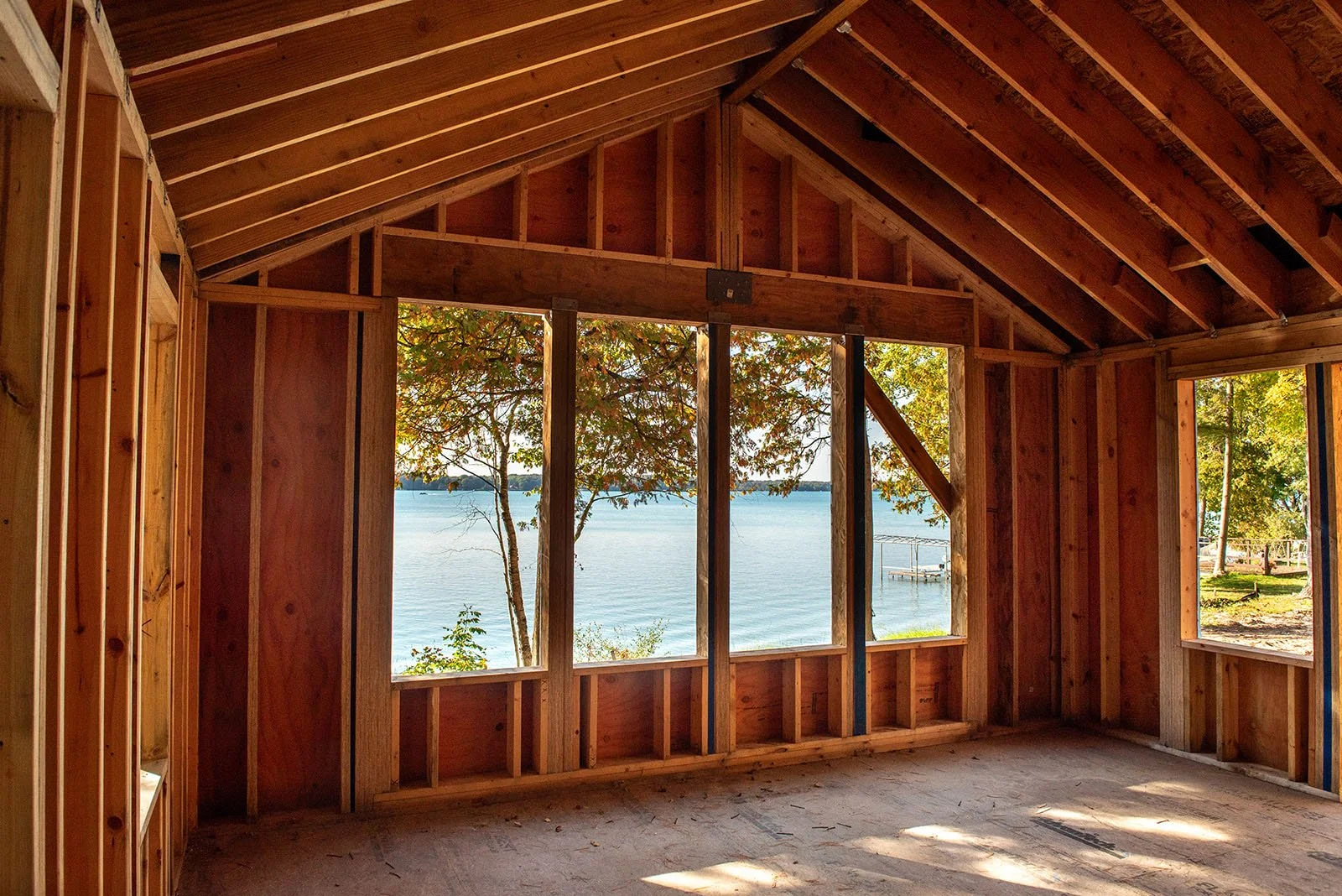 Interior view of a house under construction with wooden framing and large opening windows showing a lake and trees outside.