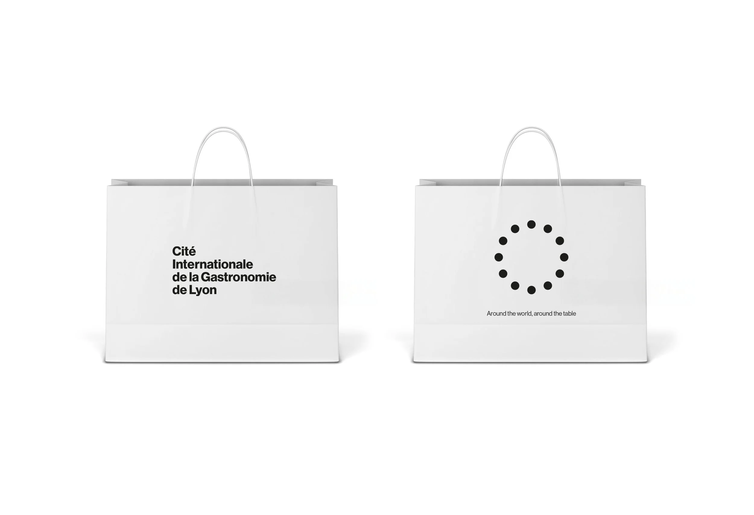 Dos bolsas de papel blancas con asas, una con texto en francés que dice 'Cité Internationale de la Gastronomie de Lyon' y otra con un círculo de puntos negros y texto en inglés 'Around the world, around the table'.