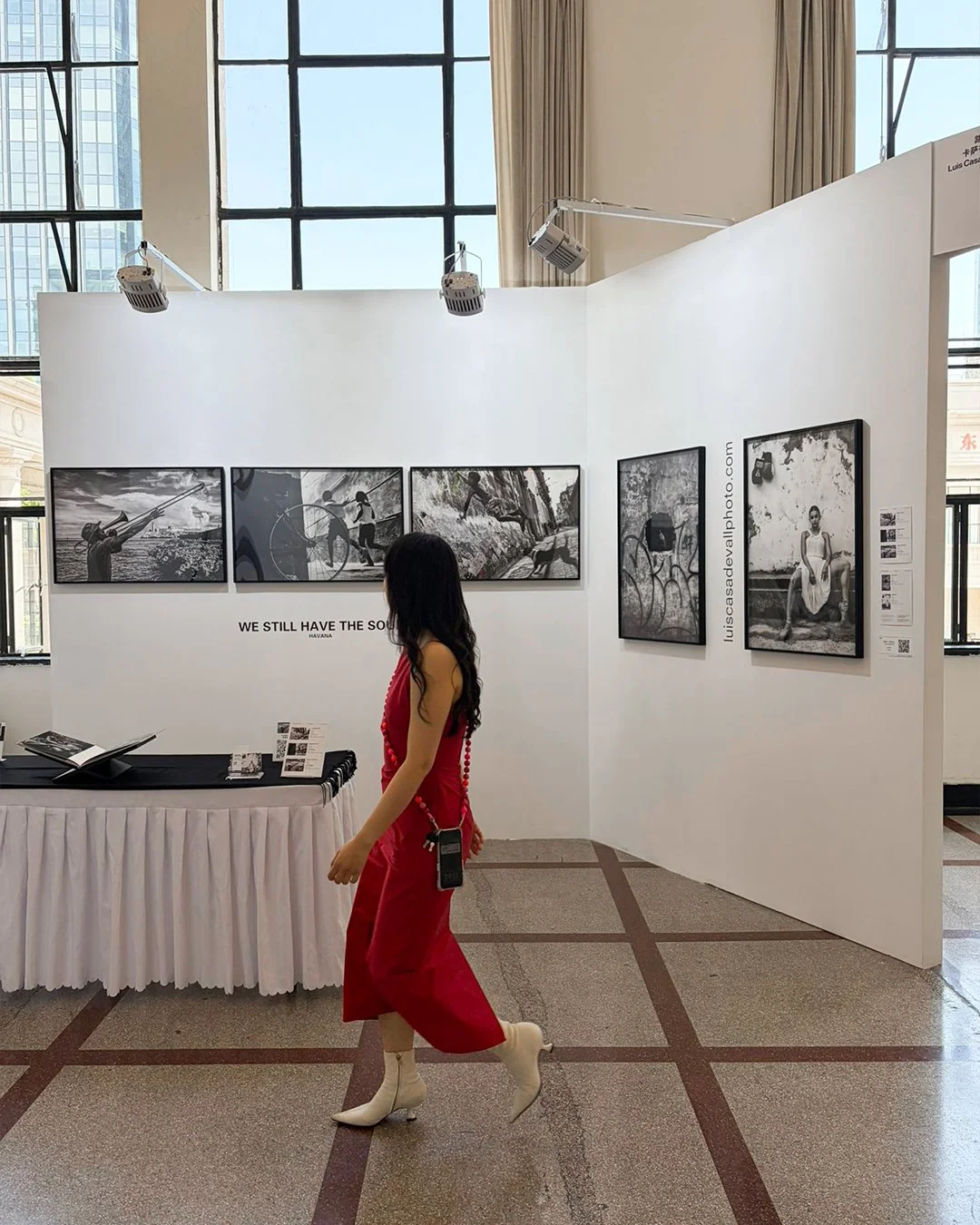 Una mujer con vestido rojo y botas blancas caminando en una galería de arte con paredes blancas y fotografías en blanco y negro exhibidas, en un ambiente con ventanas grandes y cortinas beige.