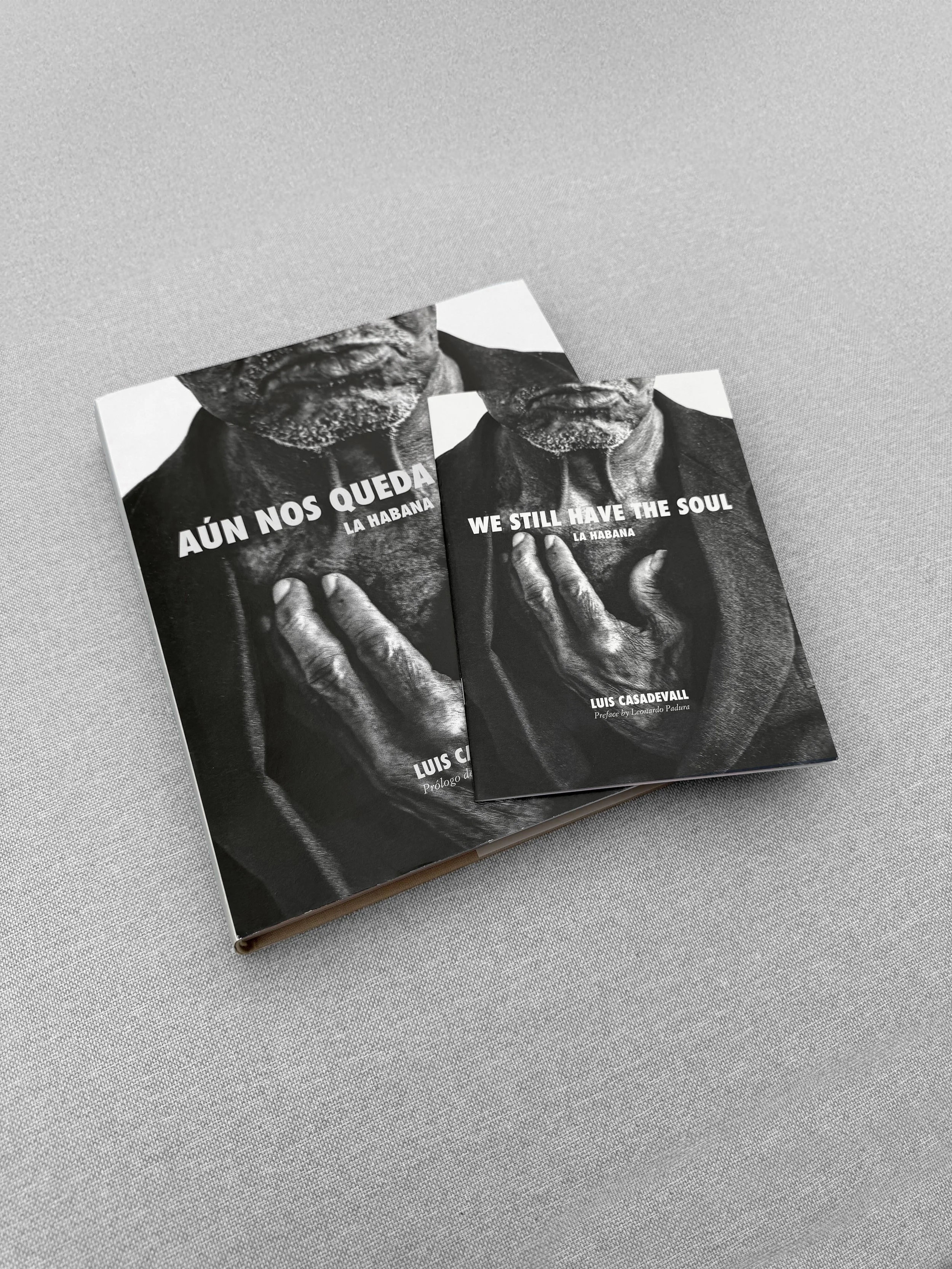 Dos libros apilados, en color negro y blanco, con una imagen de una persona mayor en la portada. Uno dice 'AÚN NOS QUEDA LA HABANA' y el otro 'WE STILL HAVE THE SOUL LA HABANA', por Luis Casadevall.