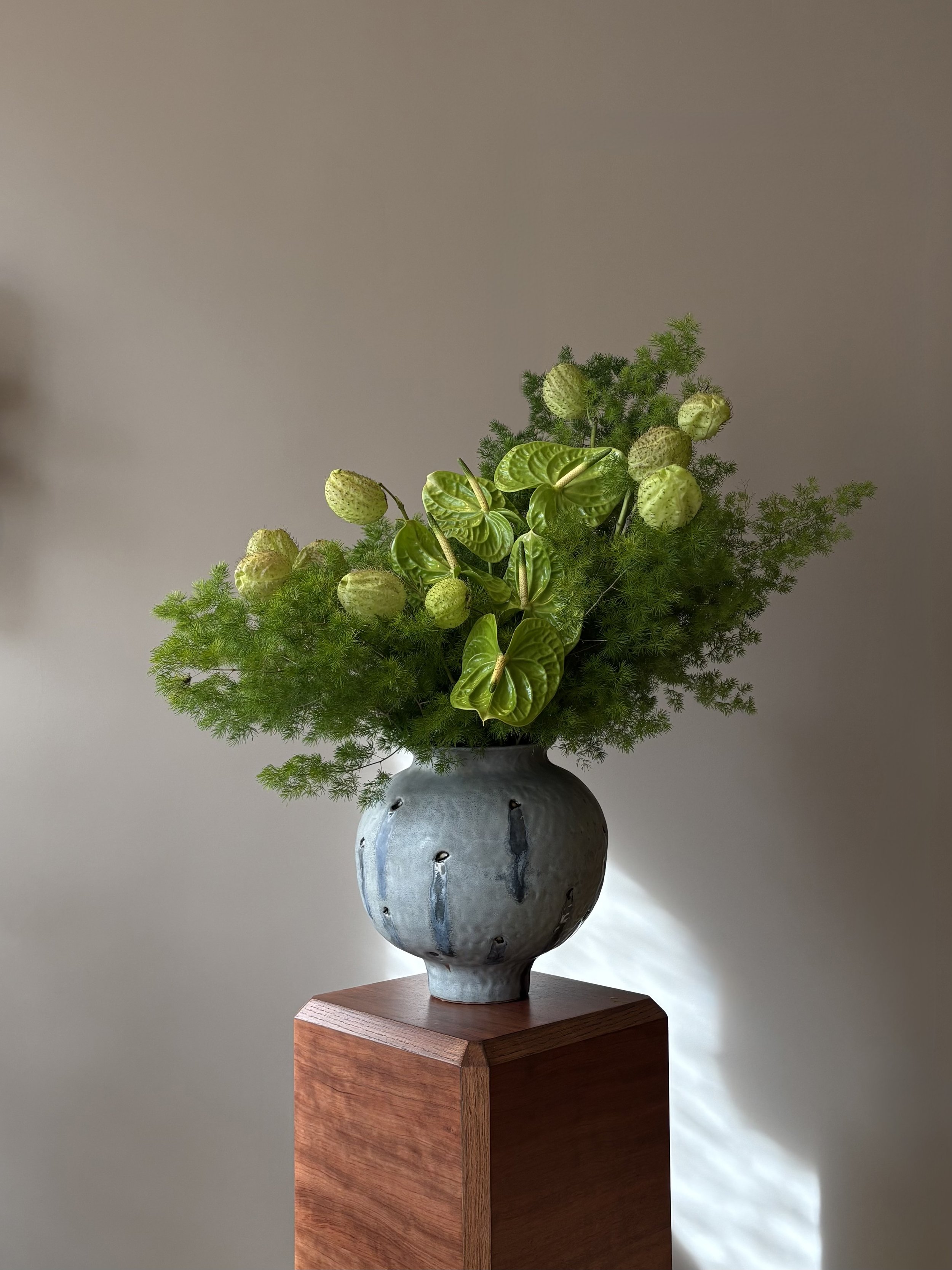 A flower arrangement in a grey ceramic vase with black accents, placed on a wooden pedestal. The arrangement includes green leafy plants and seed pods.