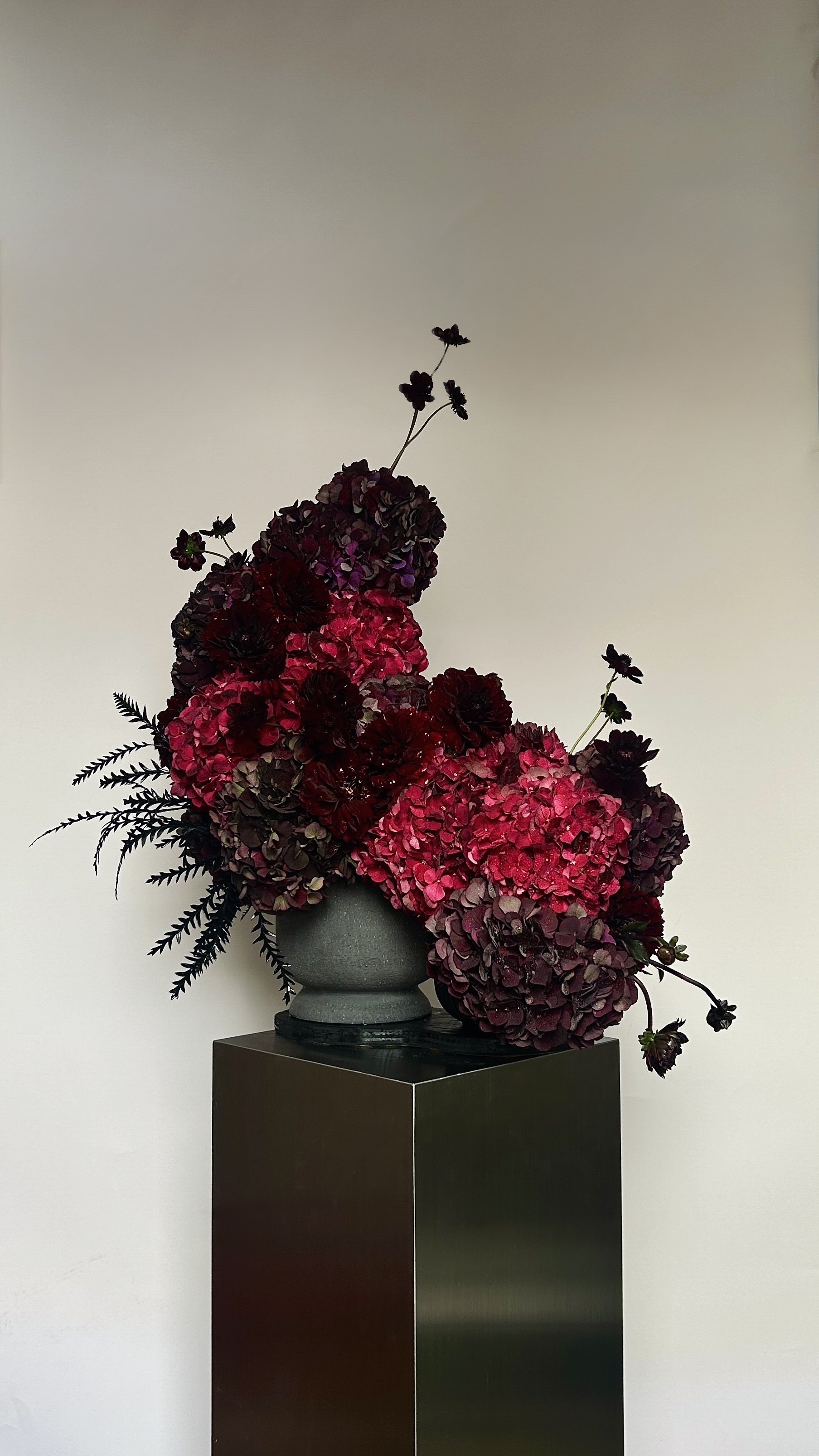 A floral arrangement with pink and dark purple hydrangeas, accented with dark foliage, in a gray vase on a black pedestal against a plain off-white wall.