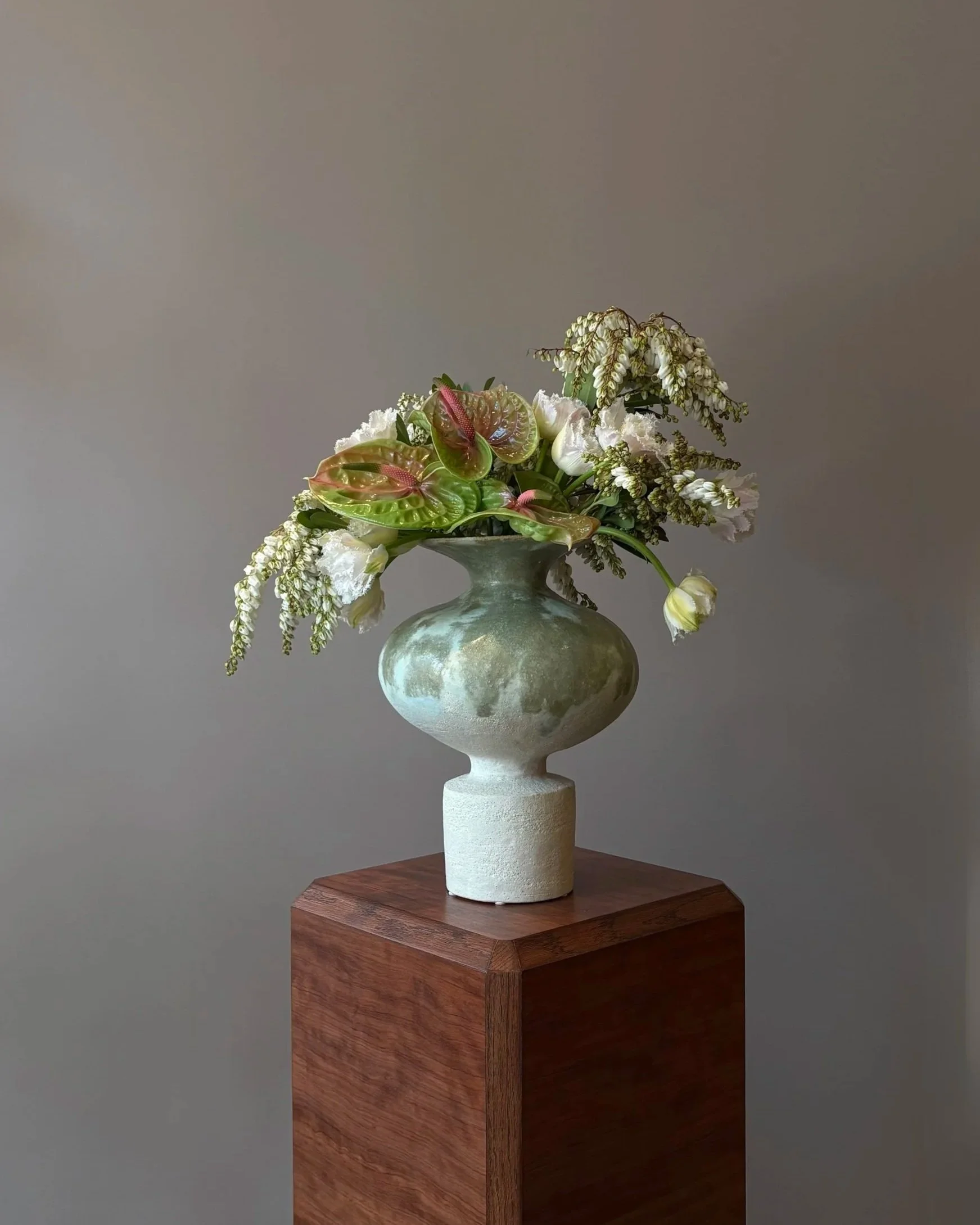 A decorative ceramic vase with a greenish glaze on a wooden pedestal, filled with white flowers, pink and green leaves, and hanging white blossoms.