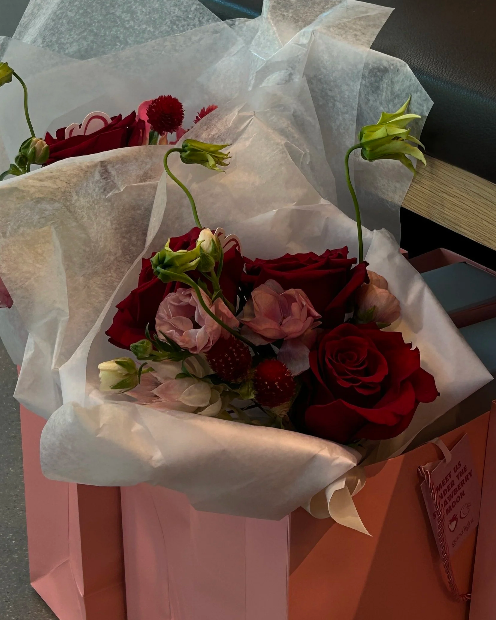 A bouquet of red roses, pink flowers, and green buds wrapped in white tissue paper and placed inside a pink gift bag.