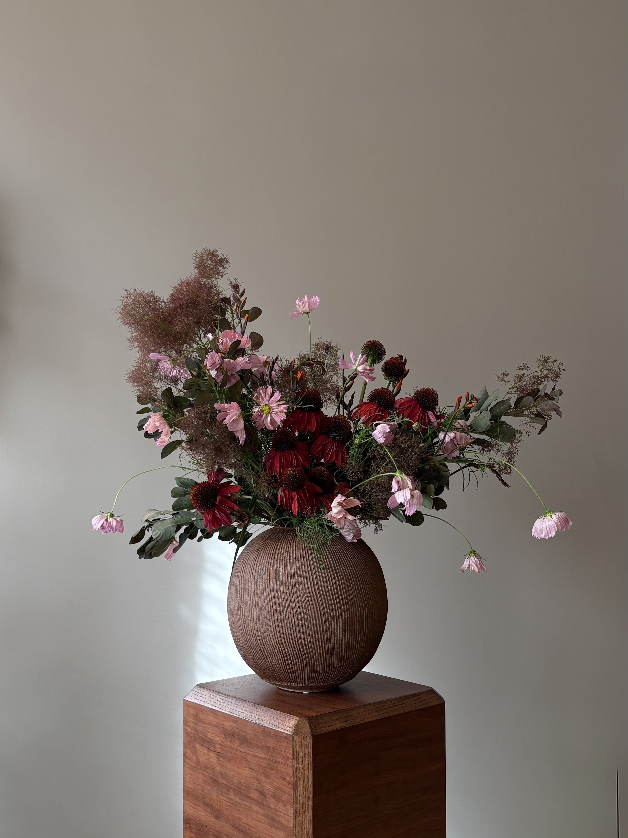 A large bouquet of pink and red flowers in a textured brown round vase on a wooden stand against a plain wall.