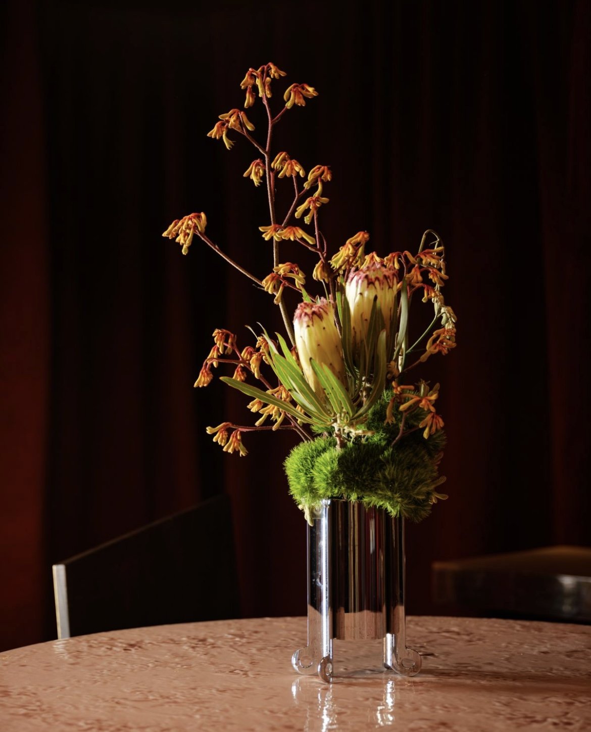 A tall, shiny silver vase containing an arrangement of exotic flowers including large, cream-colored blooms, and small yellow and orange flowers with dark orange edges, set on a polished wooden table against a dark background.