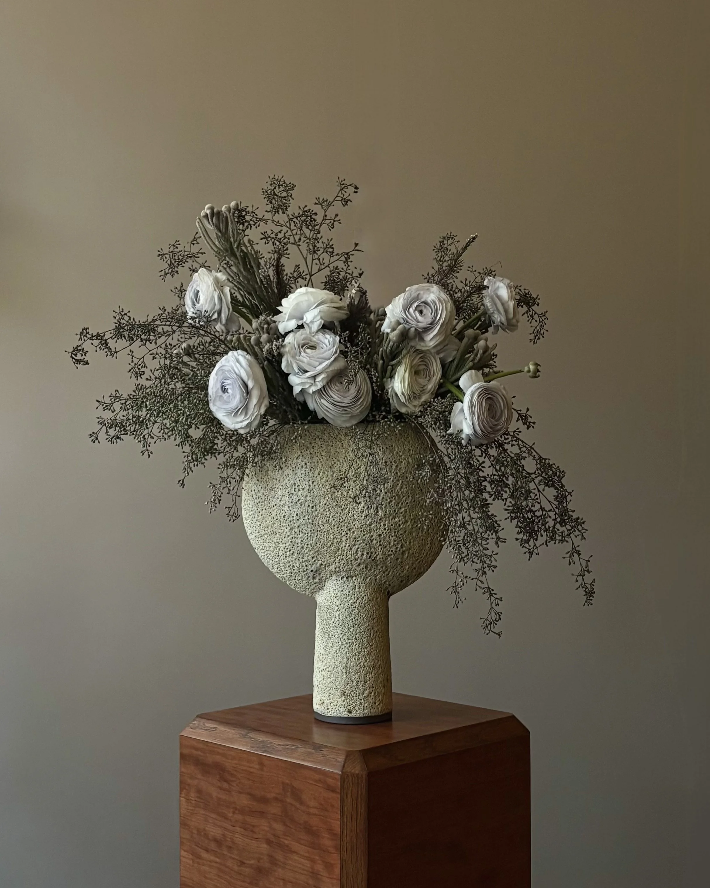 A beige textured vase with a rounded top and a narrow stem, containing white and cream-colored flowers with foliage, placed on a wooden pedestal.